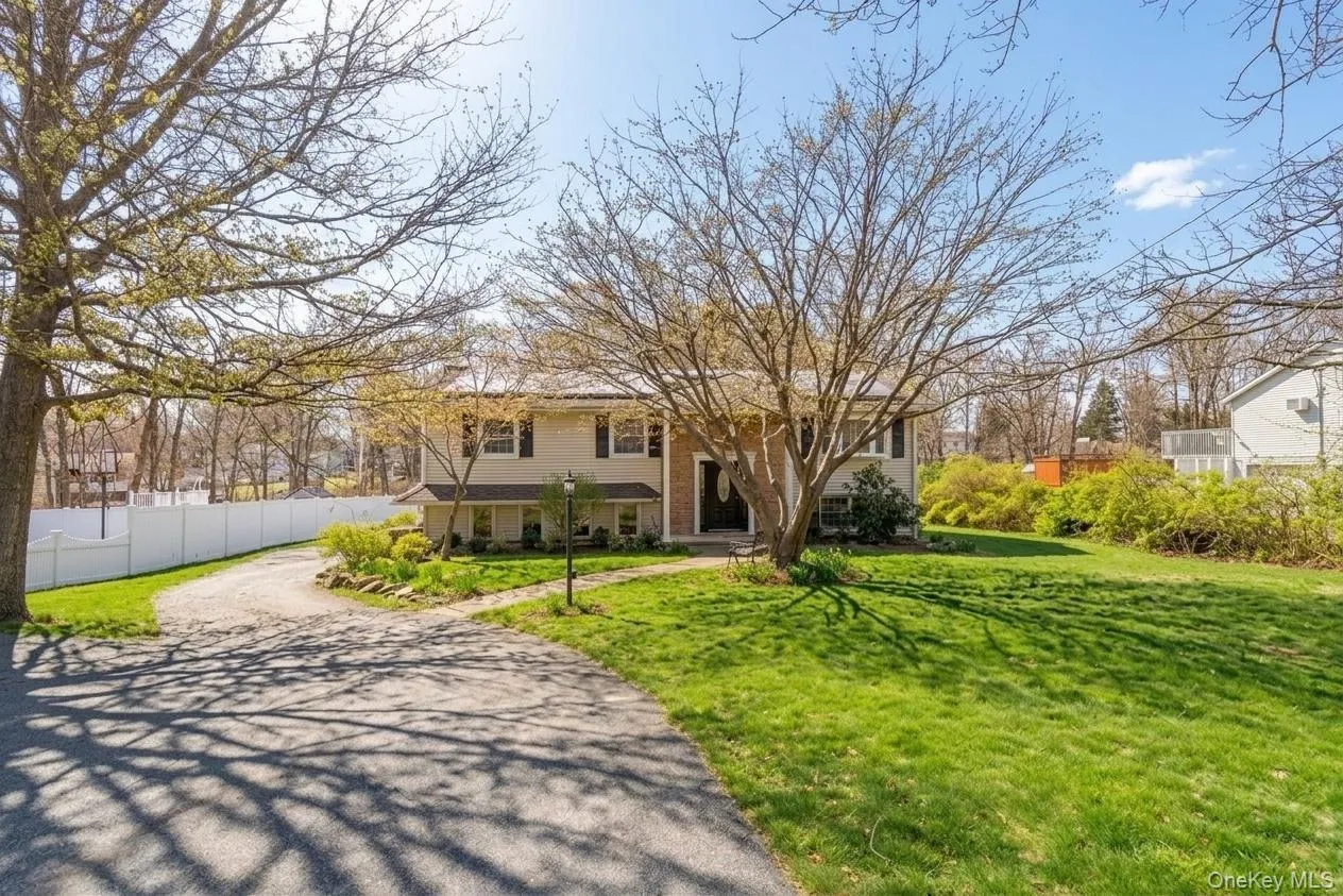 Two-story residence featuring a paved driveway, front lawn, mature trees, and a white privacy fence Two-story residence featuring a paved driveway, front lawn, mature trees, and a white privacy fence