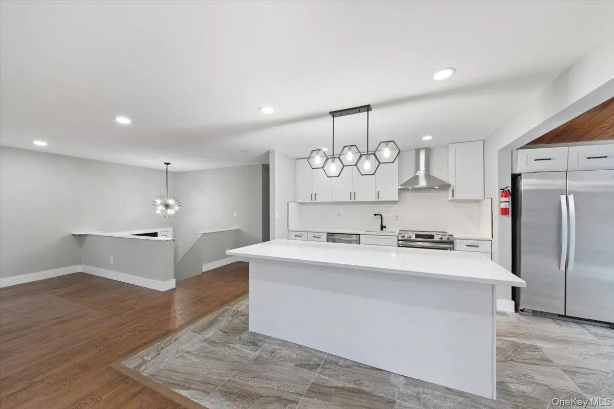 Open-concept kitchen featuring a large island with white countertop, stainless steel appliances, a geometric pendant light fixture, and light-toned cabinetry Open-concept kitchen featuring a large island with white countertop, stainless steel appliances, a geometric pendant light fixture, and light-toned cabinetry