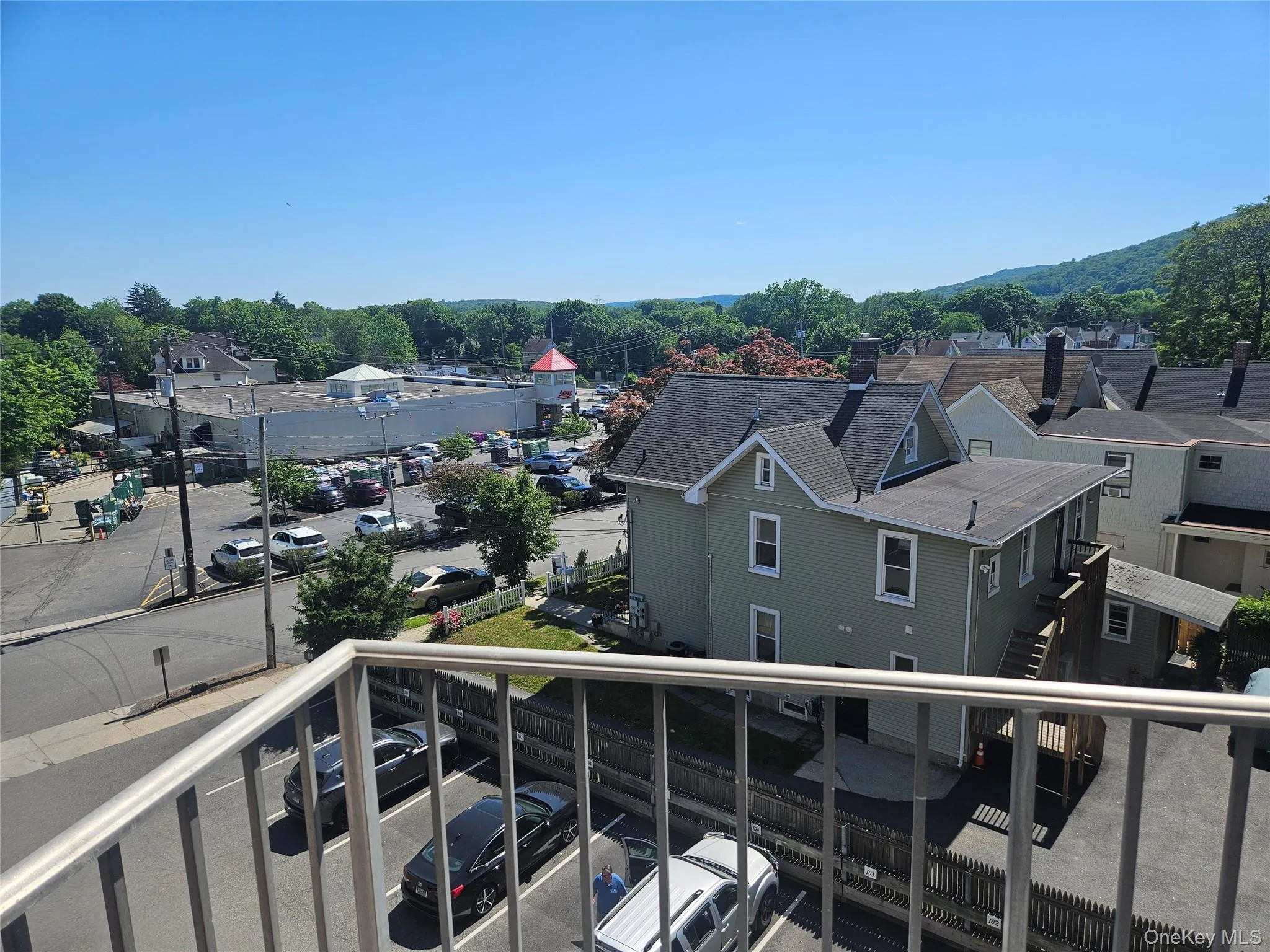 Balcony railing overlooking a parking area, surrounded by commercial buildings, residential structures, and distant mountains. Balcony railing overlooking a parking area, surrounded by commercial buildings, residential structures, and distant mountains.