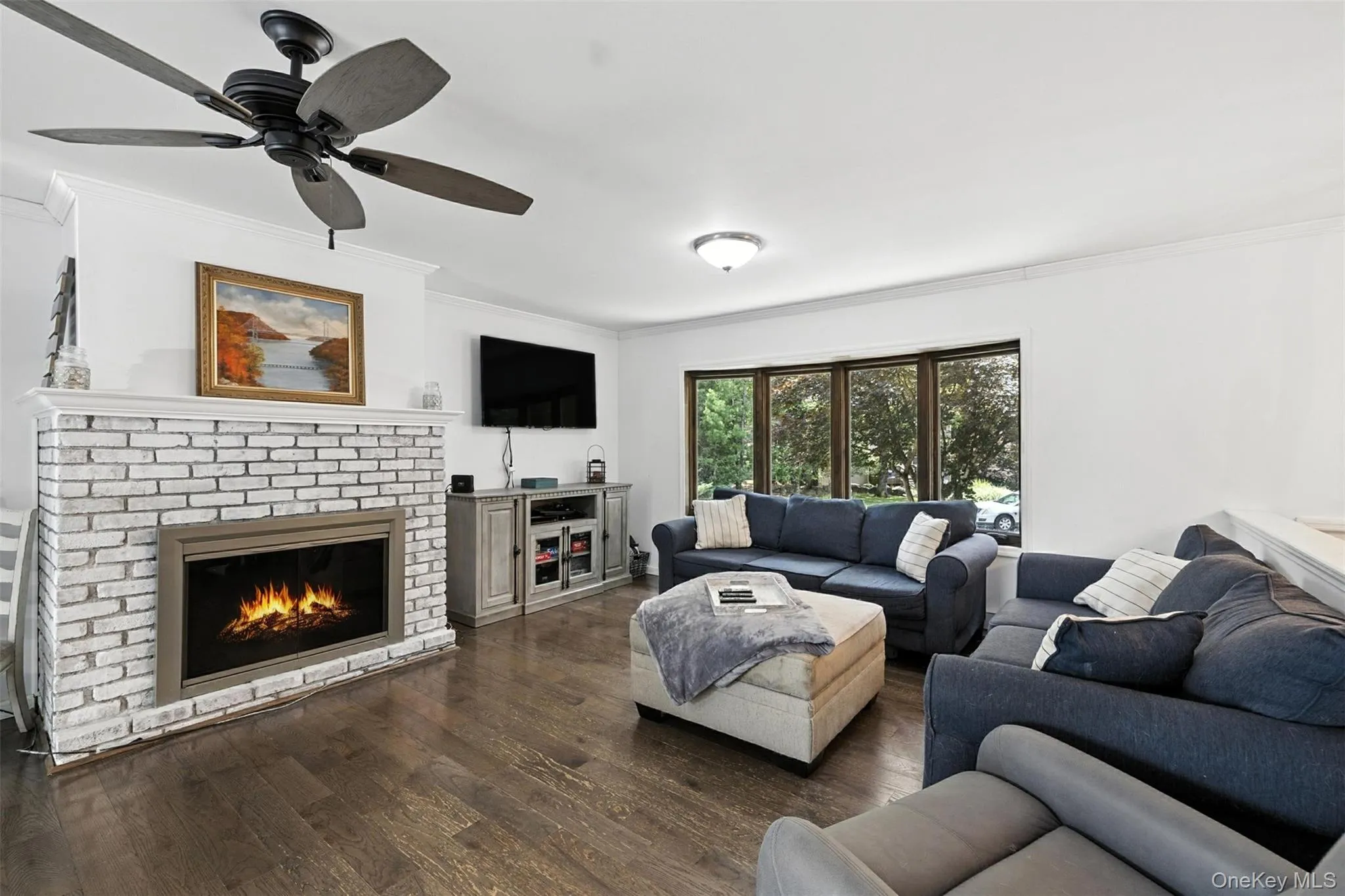 Living area featuring crown molding, dark wood-type flooring, a brick fireplace, and a ceiling fan Living area featuring crown molding, dark wood-type flooring, a brick fireplace, and a ceiling fan