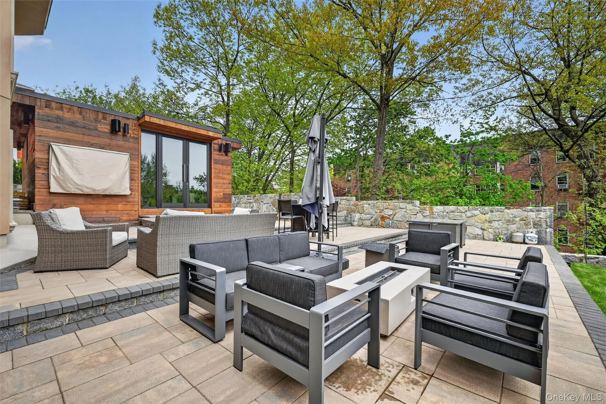 Expansive outdoor patio featuring an integrated fire pit, stone retaining walls, and a modern ancillary structure with wood paneling and large glass doors Expansive outdoor patio featuring an integrated fire pit, stone retaining walls, and a modern ancillary structure with wood paneling and large glass doors