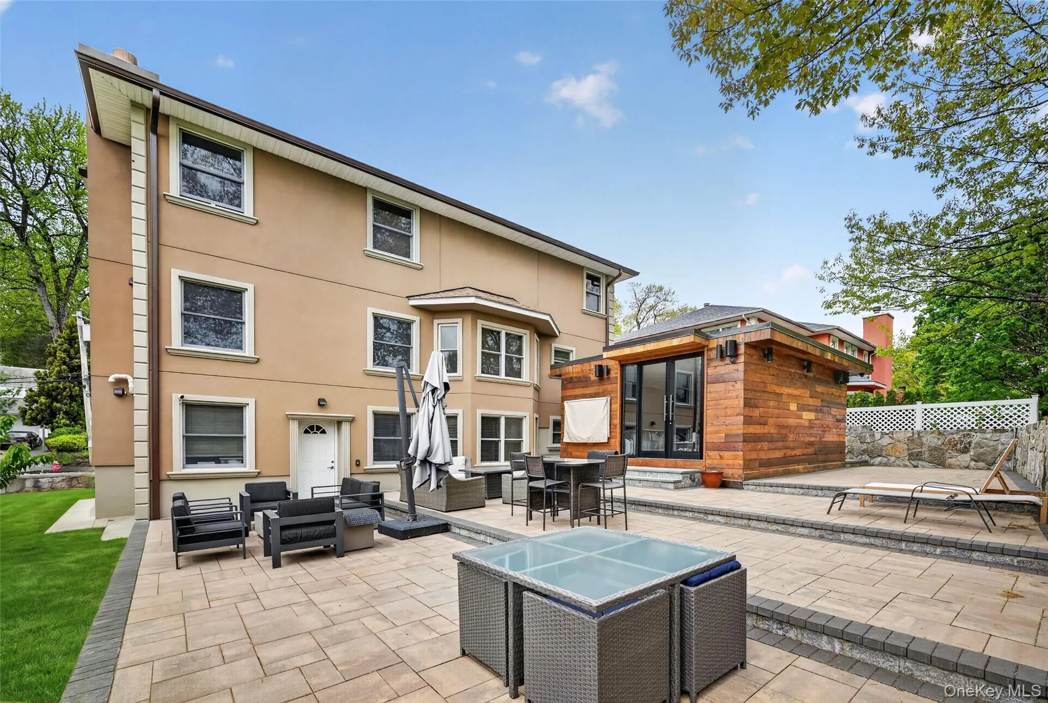 Expansive paver patio featuring multiple seating areas, a separate wood-paneled structure with large glass doors, and a stone retaining wall Expansive paver patio featuring multiple seating areas, a separate wood-paneled structure with large glass doors, and a stone retaining wall