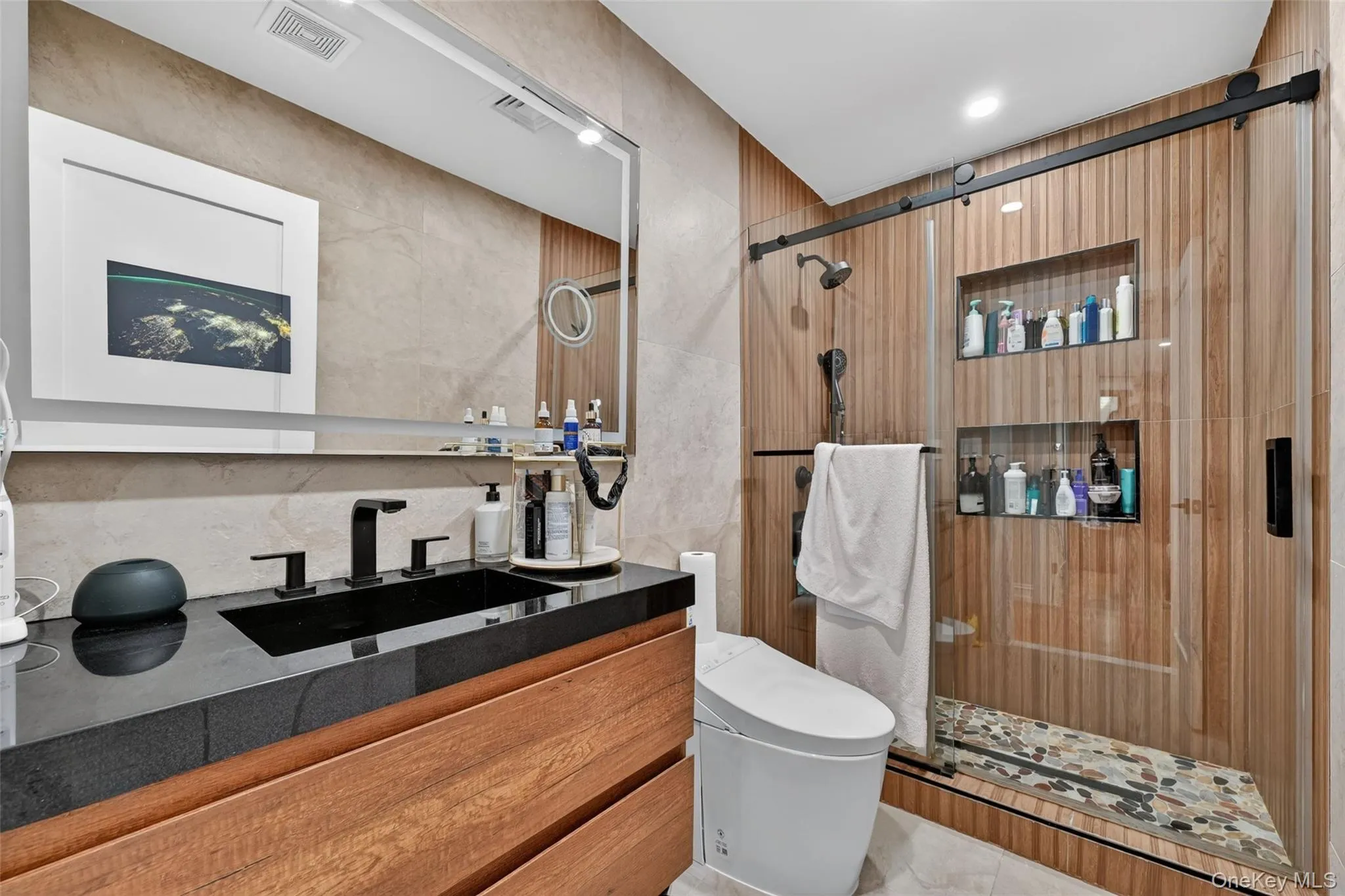Bathroom featuring a wood-finish vanity with a black countertop and integrated sink, a large wall-mounted mirror, and a glass-enclosed shower Bathroom featuring a wood-finish vanity with a black countertop and integrated sink, a large wall-mounted mirror, and a glass-enclosed shower