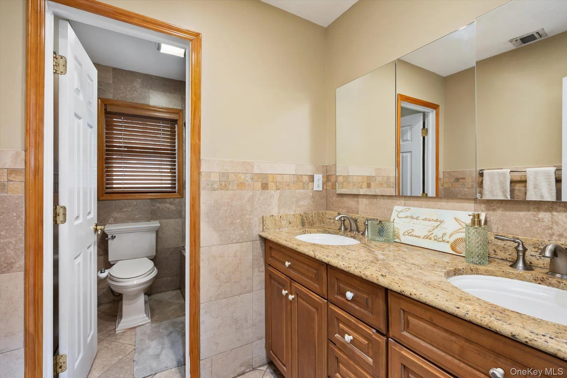 Full bath featuring double vanity, a wainscoted wall, light tile patterned floors, and tile walls Full bath featuring double vanity, a wainscoted wall, light tile patterned floors, and tile walls