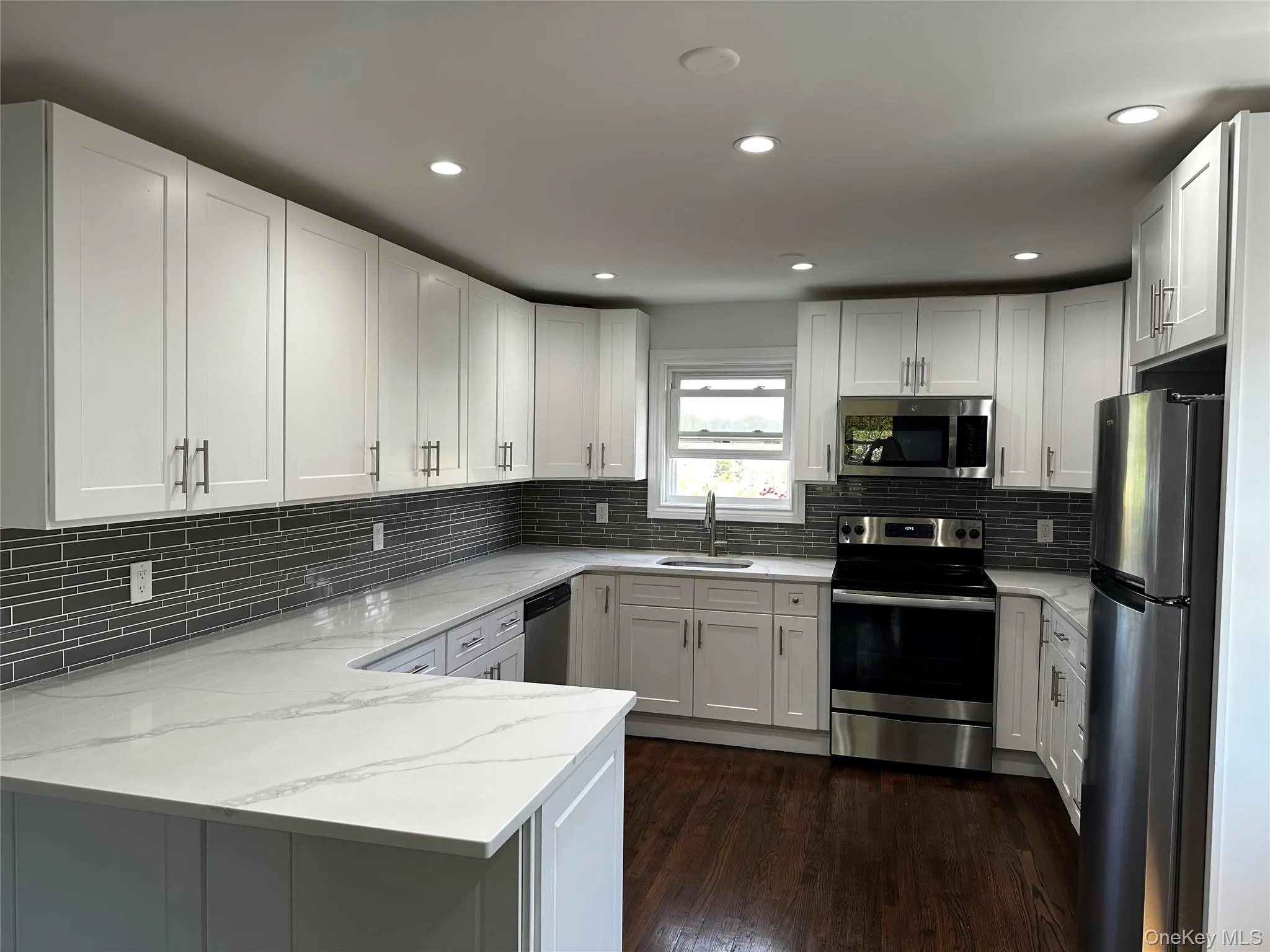 Bright kitchen featuring white shaker cabinetry, stainless steel appliances, white patterned countertops, a gray mosaic tile backsplash, and dark wood-finish flooring Bright kitchen featuring white shaker cabinetry, stainless steel appliances, white patterned countertops, a gray mosaic tile backsplash, and dark wood-finish flooring