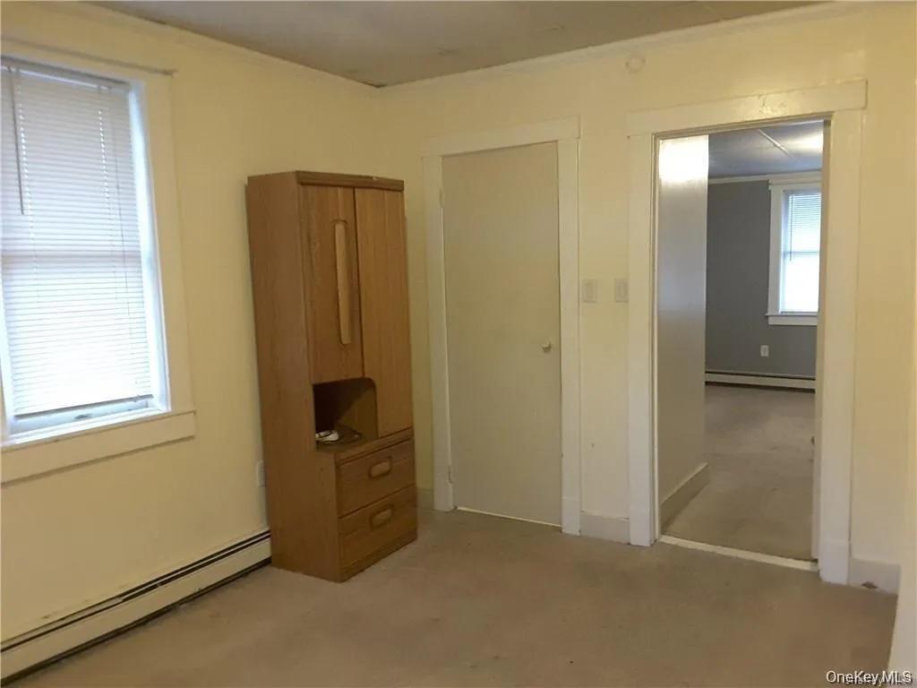 Carpeted interior room with white trim, baseboard heating, a single window with blinds, and a white interior door Carpeted interior room with white trim, baseboard heating, a single window with blinds, and a white interior door