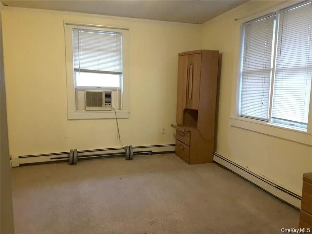 Carpeted room featuring multiple windows with blinds, a wall-mounted air conditioning unit, and baseboard heating Carpeted room featuring multiple windows with blinds, a wall-mounted air conditioning unit, and baseboard heating
