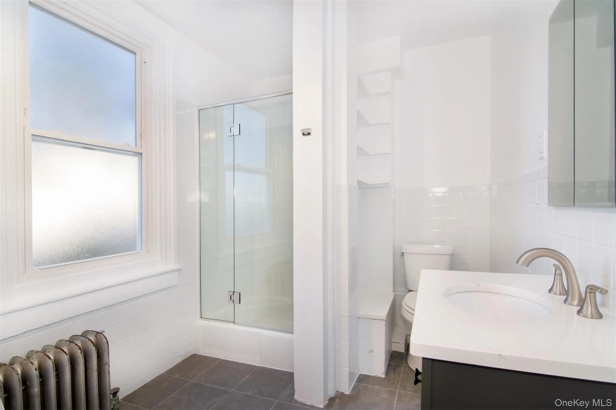 Full bathroom with radiator heating unit, tile patterned flooring, shower with glass door, toilet, and tile walls Full bathroom with radiator heating unit, tile patterned flooring, shower with glass door, toilet, and tile walls