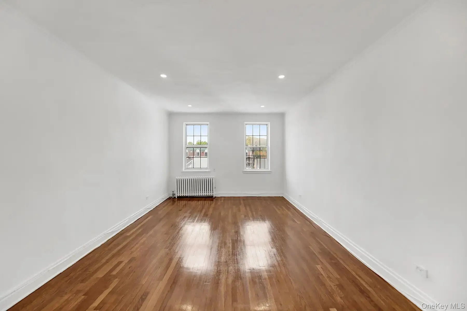 Spacious room featuring hardwood flooring, white walls, two double-hung windows, recessed lighting, and a radiator Spacious room featuring hardwood flooring, white walls, two double-hung windows, recessed lighting, and a radiator