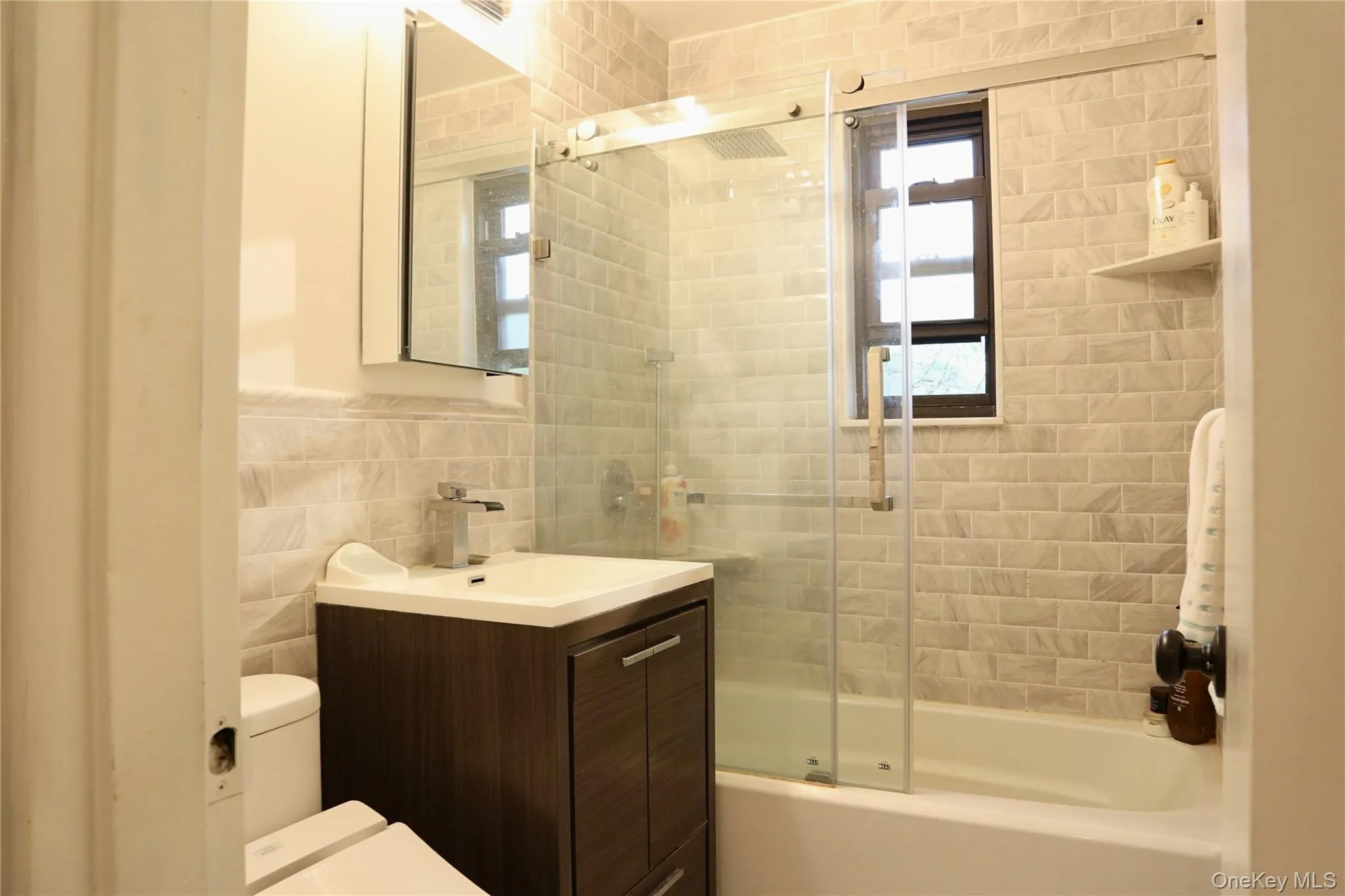 Bathroom featuring a modern vanity with integrated sink, glass-enclosed shower over tub, tiled walls with a subway pattern, and a window Bathroom featuring a modern vanity with integrated sink, glass-enclosed shower over tub, tiled walls with a subway pattern, and a window