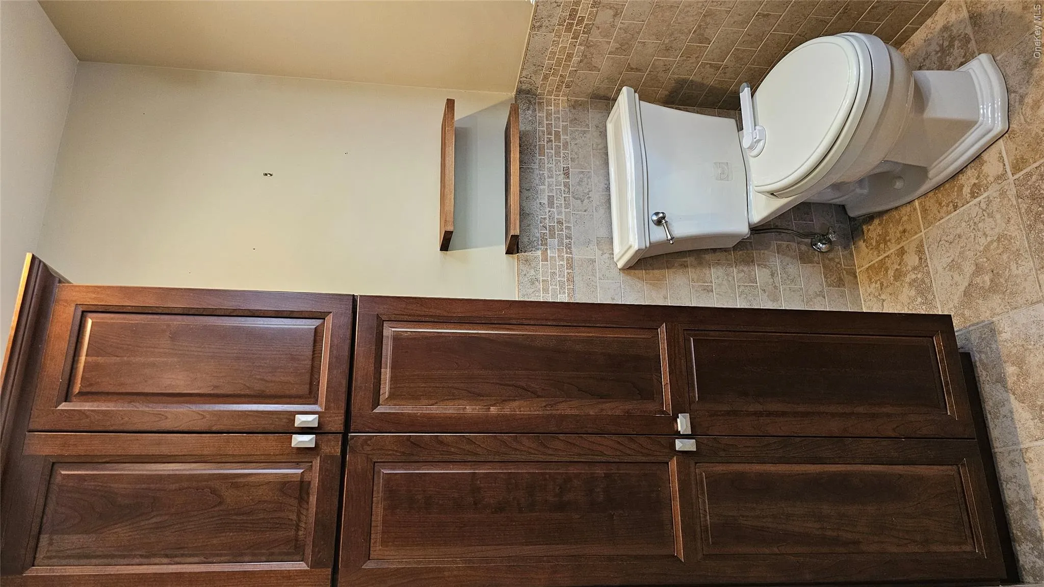 Tall wood-finish cabinetry providing ample storage, complemented by a white toilet, mosaic tile wainscoting, two floating wood-finish shelves, and light-toned floor tiles Tall wood-finish cabinetry providing ample storage, complemented by a white toilet, mosaic tile wainscoting, two floating wood-finish shelves, and light-toned floor tiles