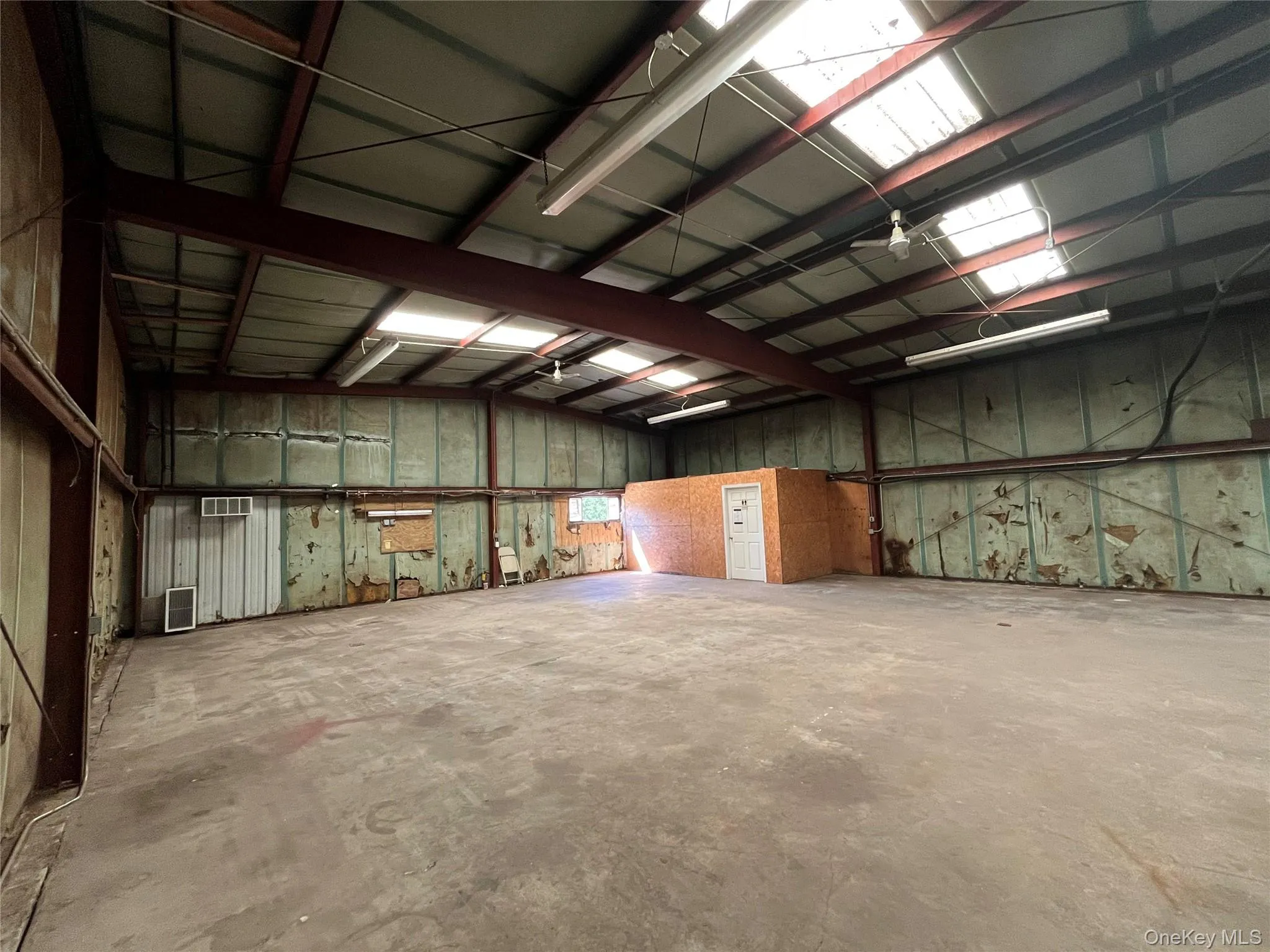 Spacious warehouse interior featuring exposed steel beams, concrete flooring, fluorescent lighting, multiple skylights, and a built-in office structure with a white door Spacious warehouse interior featuring exposed steel beams, concrete flooring, fluorescent lighting, multiple skylights, and a built-in office structure with a white door