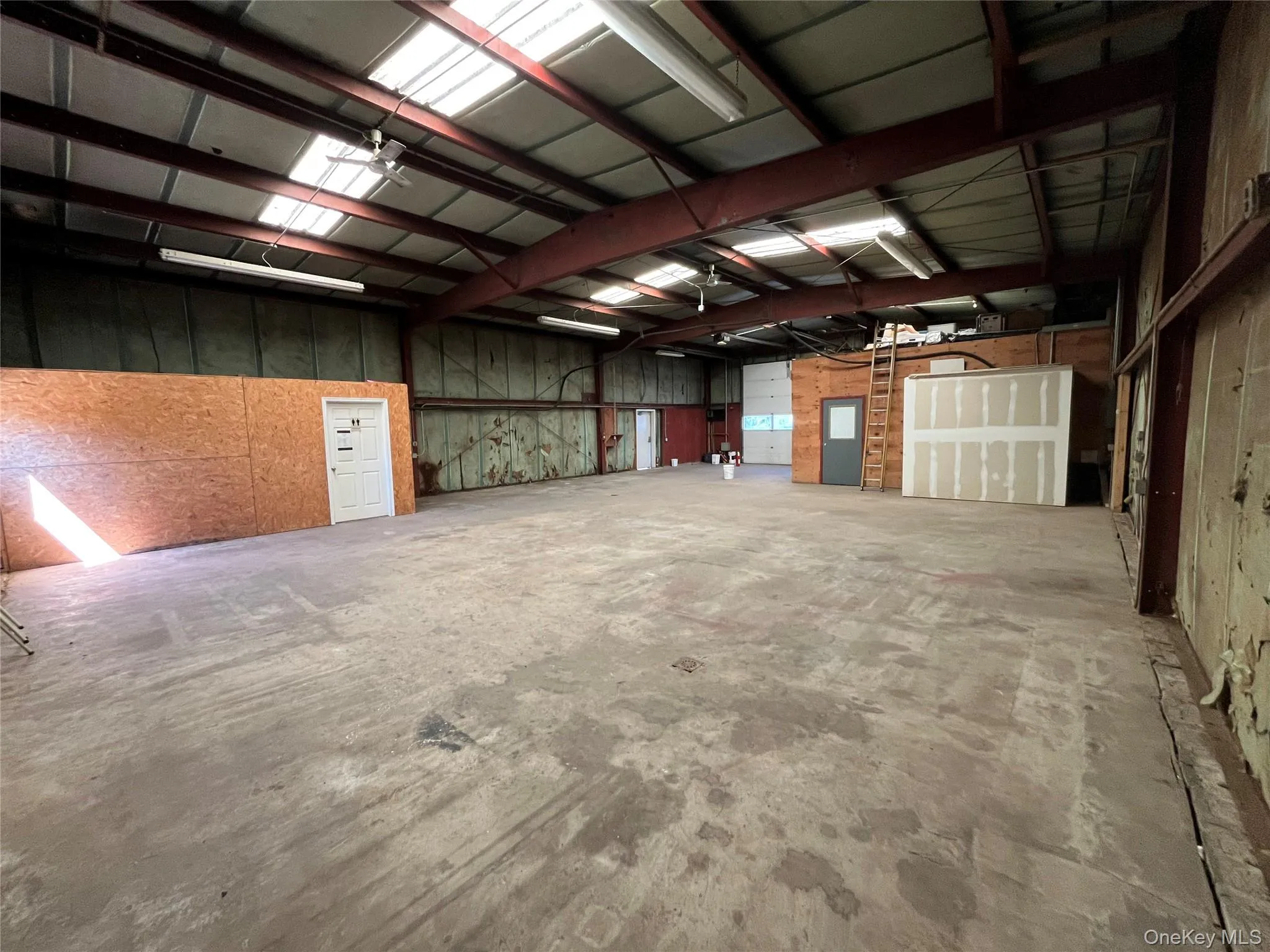 Spacious industrial interior featuring a high ceiling, exposed steel beams, concrete flooring, and multiple overhead light fixtures Spacious industrial interior featuring a high ceiling, exposed steel beams, concrete flooring, and multiple overhead light fixtures