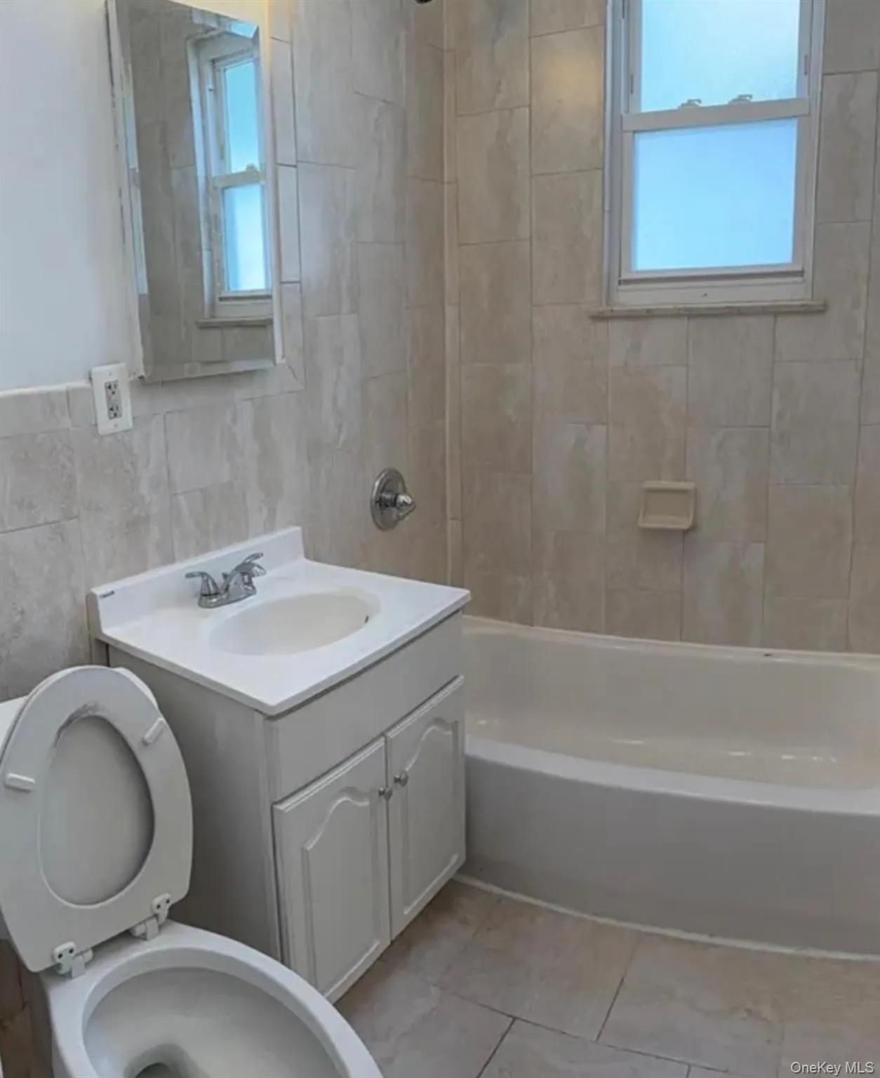 Bathroom featuring light-toned tile flooring and walls, white vanity with integrated sink, white bathtub, and a window with privacy glass Bathroom featuring light-toned tile flooring and walls, white vanity with integrated sink, white bathtub, and a window with privacy glass