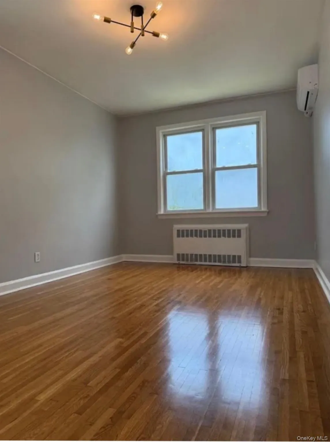 Room featuring wood flooring, a modern sputnik-style ceiling light fixture, two pane windows, a wall-mounted heating/cooling unit, and a radiator Room featuring wood flooring, a modern sputnik-style ceiling light fixture, two pane windows, a wall-mounted heating/cooling unit, and a radiator