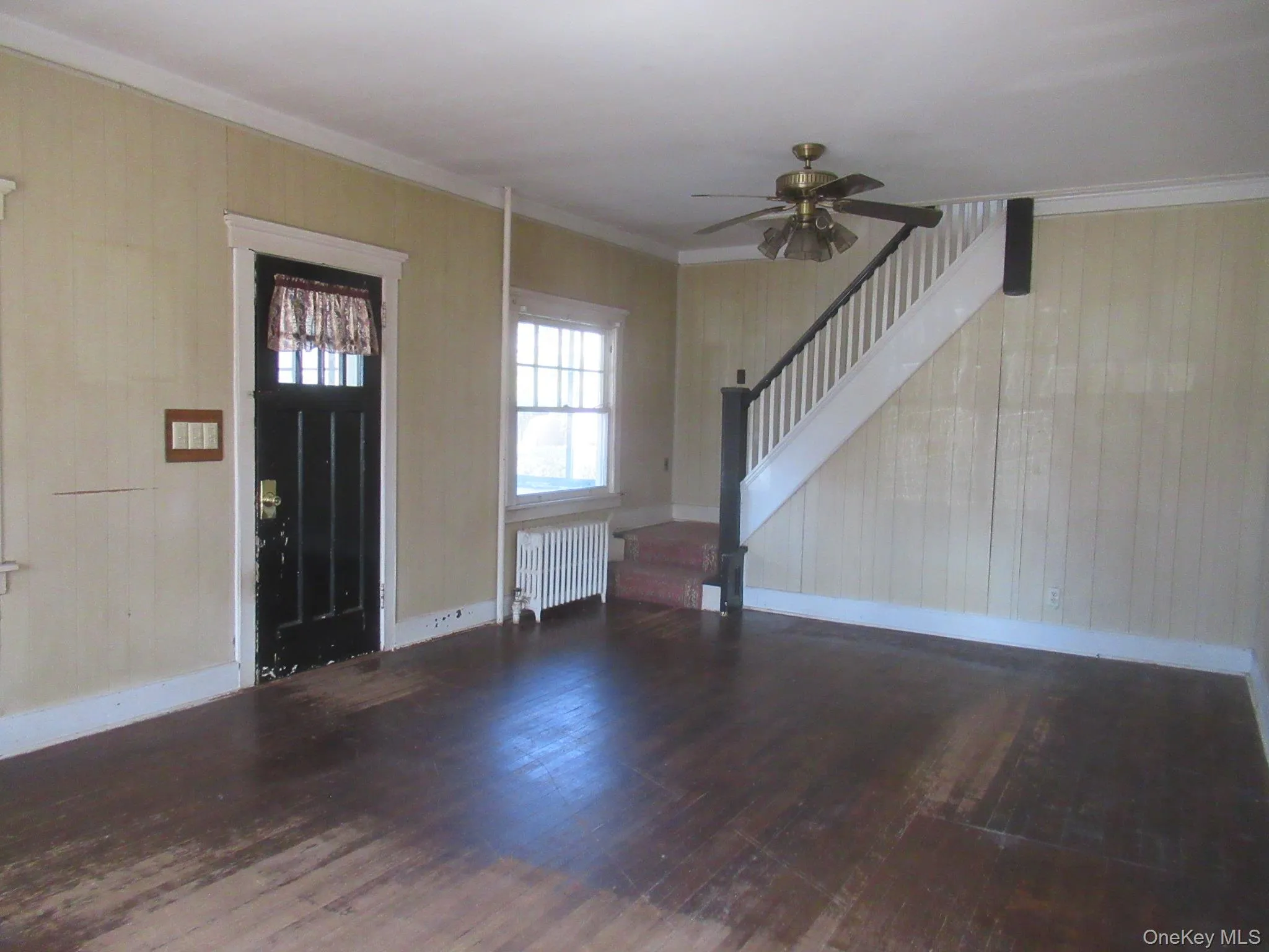 Living area featuring wood-finish flooring, a staircase with white balusters and a dark newel post, a ceiling fan, a window with a radiator below, and a multi-pane entry door Living area featuring wood-finish flooring, a staircase with white balusters and a dark newel post, a ceiling fan, a window with a radiator below, and a multi-pane entry door