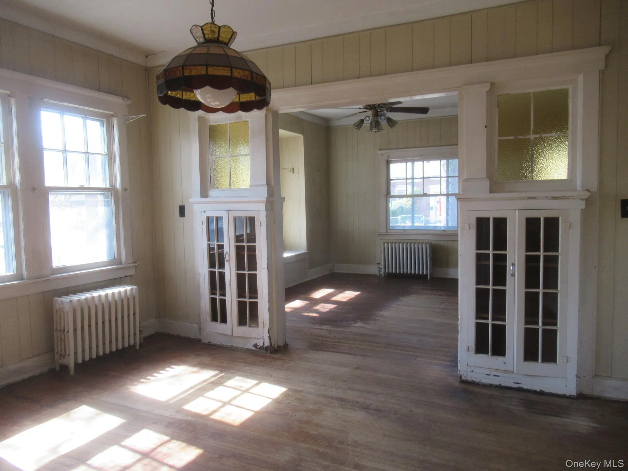 Interior space featuring wood-finish flooring, multiple windows, built-in cabinetry with glass panel doors, and a decorative overhead light fixture Interior space featuring wood-finish flooring, multiple windows, built-in cabinetry with glass panel doors, and a decorative overhead light fixture