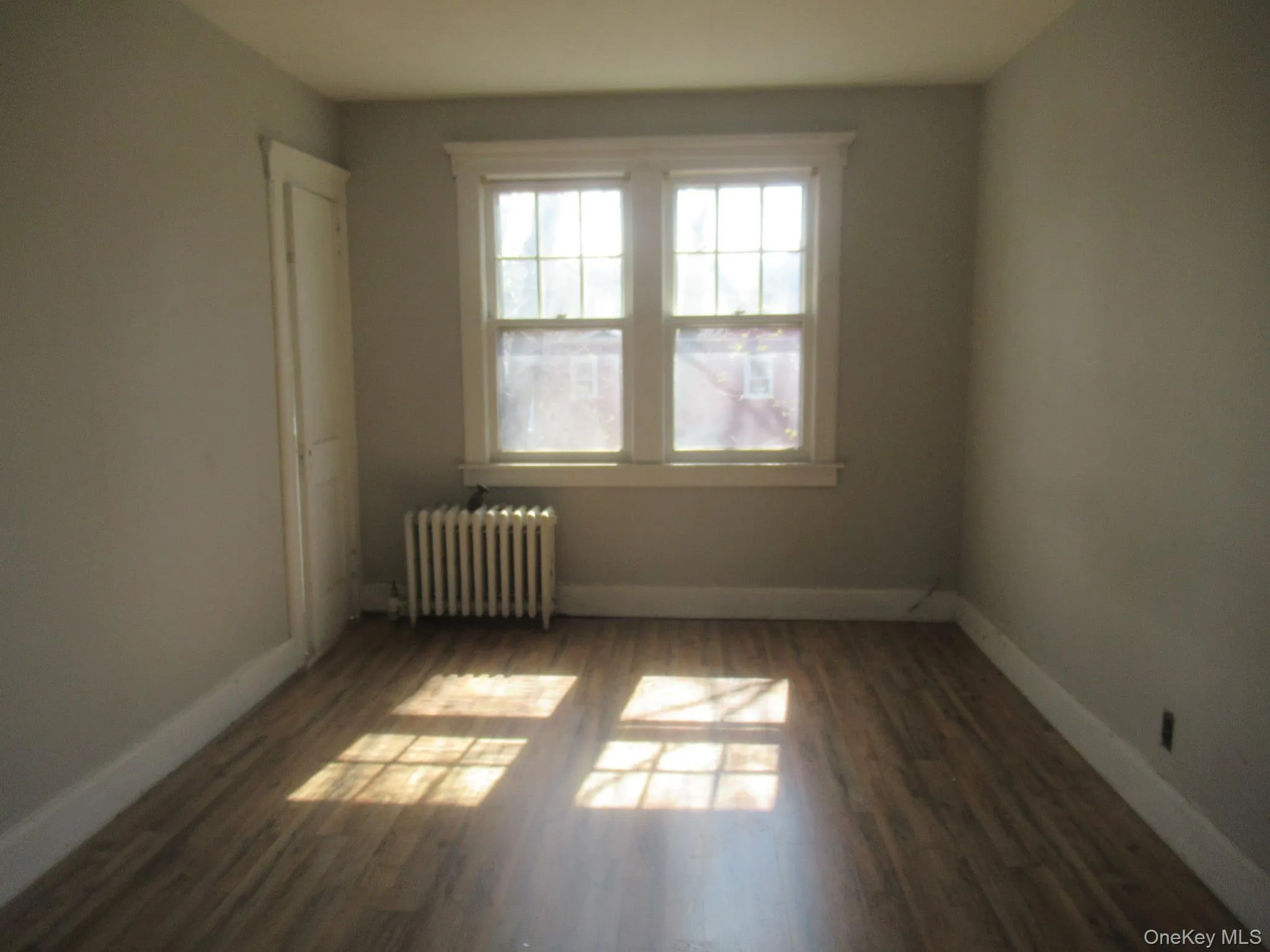 Room featuring wood-finish flooring, a double window with white trim, and a wall-mounted radiator Room featuring wood-finish flooring, a double window with white trim, and a wall-mounted radiator