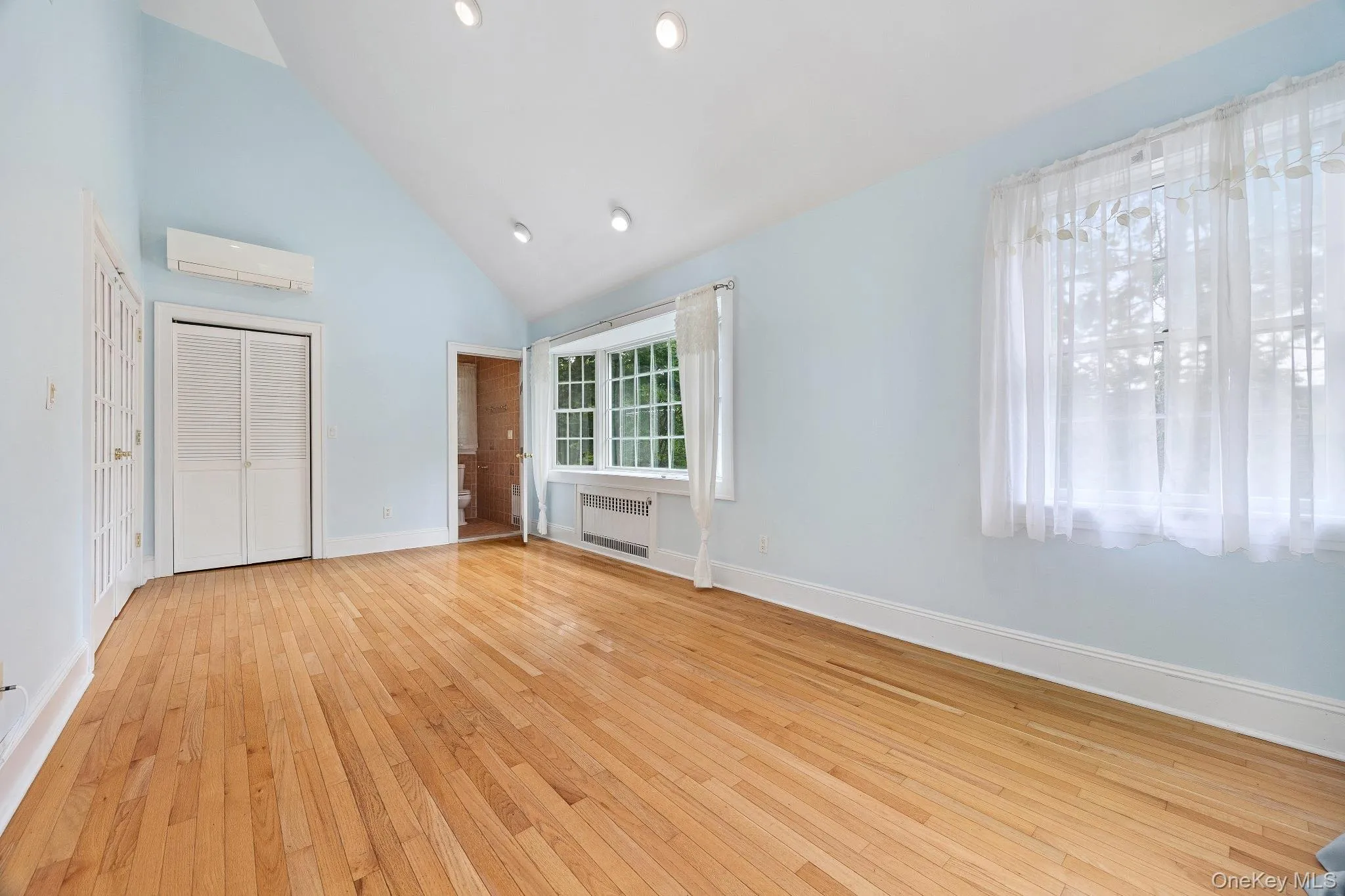 Unfurnished bedroom featuring radiator heating unit, a wall mounted air conditioner, high vaulted ceiling, light wood-style floors, and a closet Unfurnished bedroom featuring radiator heating unit, a wall mounted air conditioner, high vaulted ceiling, light wood-style floors, and a closet