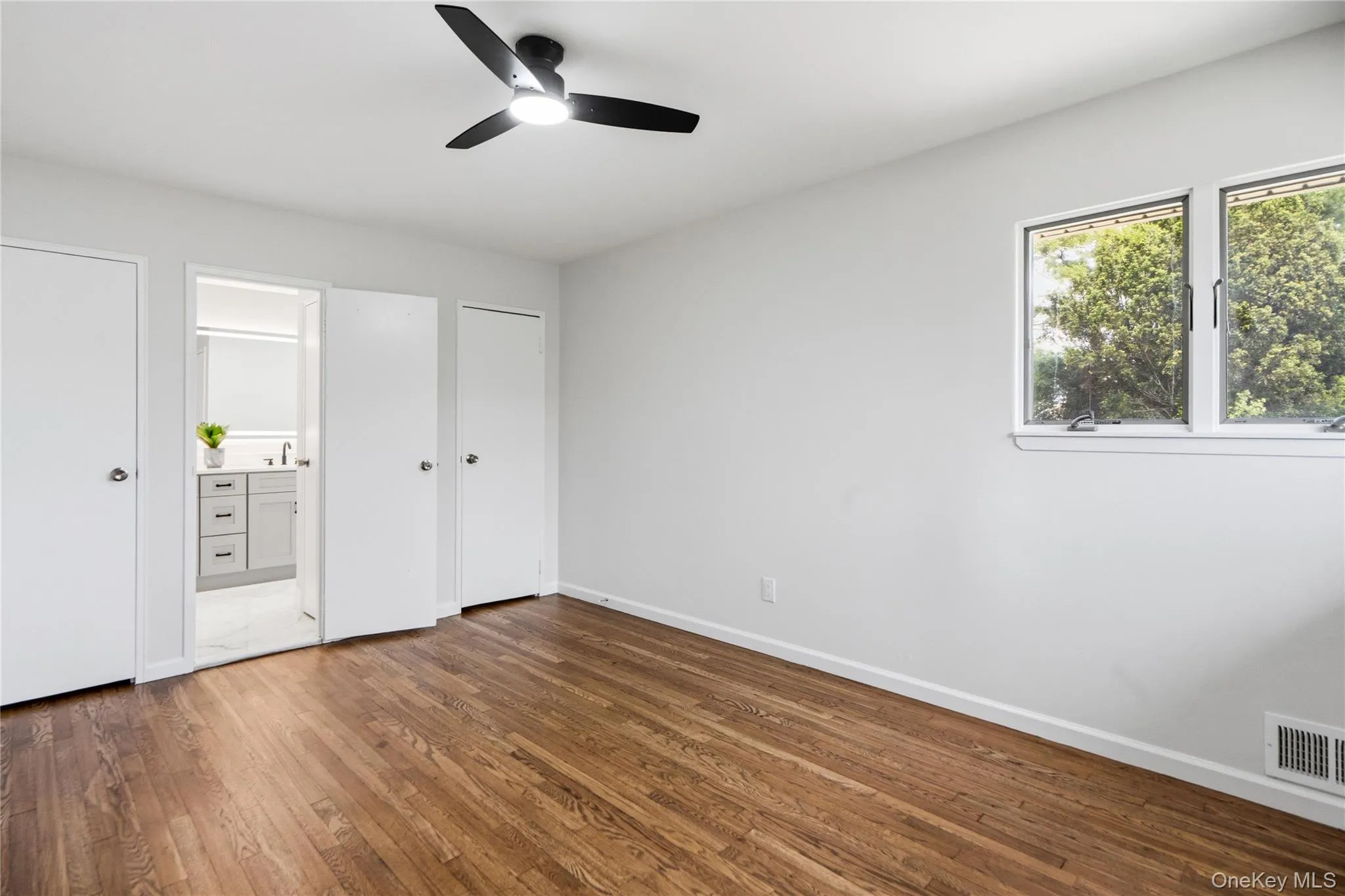 Spacious room featuring wood-finish flooring, a modern black ceiling fan with integrated lighting, and two windows providing natural light Spacious room featuring wood-finish flooring, a modern black ceiling fan with integrated lighting, and two windows providing natural light