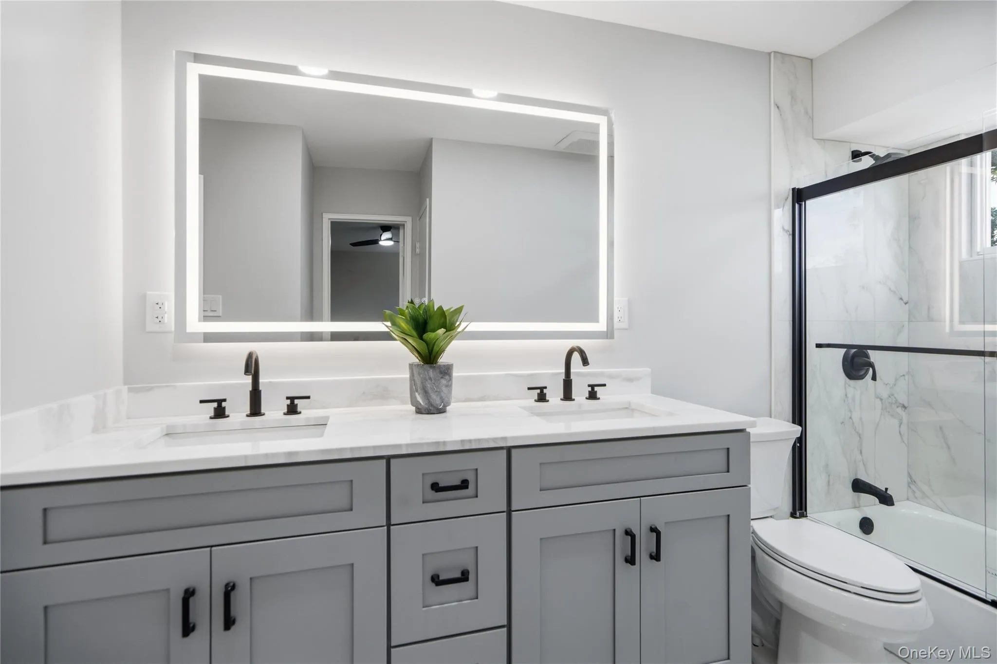 Bathroom featuring a dual vanity with a light gray cabinet and white quartz countertop, backlit mirror, matte black hardware, and a tub-shower combination with a clear glass enclosure and white marble-look tile surround Bathroom featuring a dual vanity with a light gray cabinet and white quartz countertop, backlit mirror, matte black hardware, and a tub-shower combination with a clear glass enclosure and white marble-look tile surround