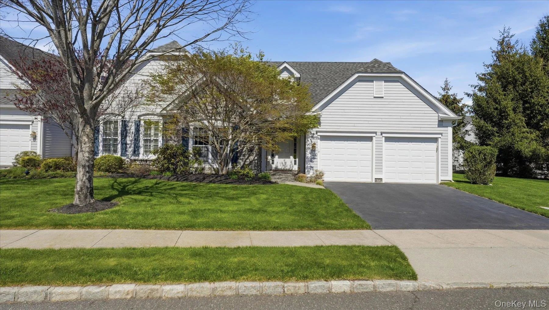 Light-colored siding exterior featuring a dark gray shingle roof, twin two-car garages, a paved driveway, and manicured front lawn with mature trees and shrubbery Light-colored siding exterior featuring a dark gray shingle roof, twin two-car garages, a paved driveway, and manicured front lawn with mature trees and shrubbery