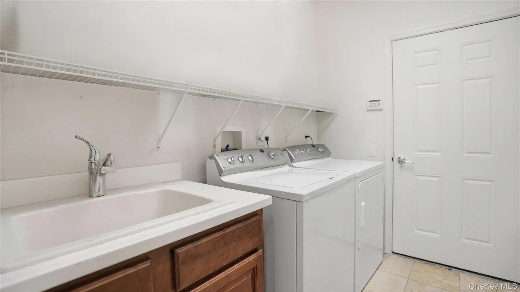Dedicated laundry area featuring a utility sink with a chrome faucet, stone-finish countertop, wood-finish cabinetry, wire shelving, and tiled flooring Dedicated laundry area featuring a utility sink with a chrome faucet, stone-finish countertop, wood-finish cabinetry, wire shelving, and tiled flooring