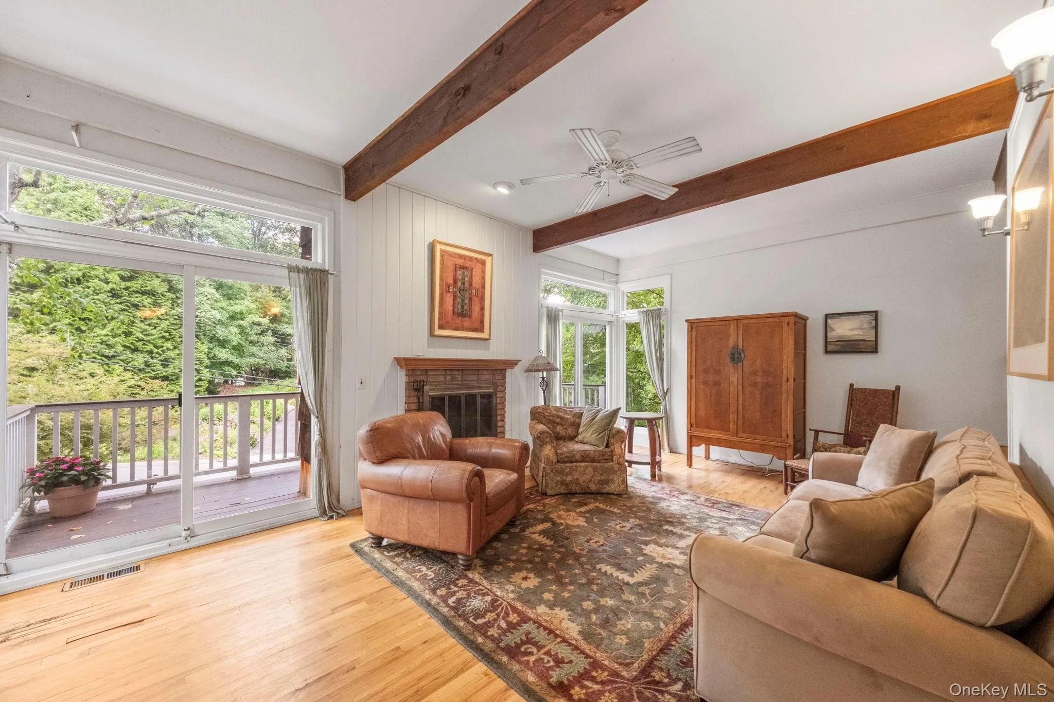 Spacious living area featuring exposed wood beams, a brick fireplace with a wood mantle, wood-finish flooring, and sliding glass doors opening to a deck Spacious living area featuring exposed wood beams, a brick fireplace with a wood mantle, wood-finish flooring, and sliding glass doors opening to a deck