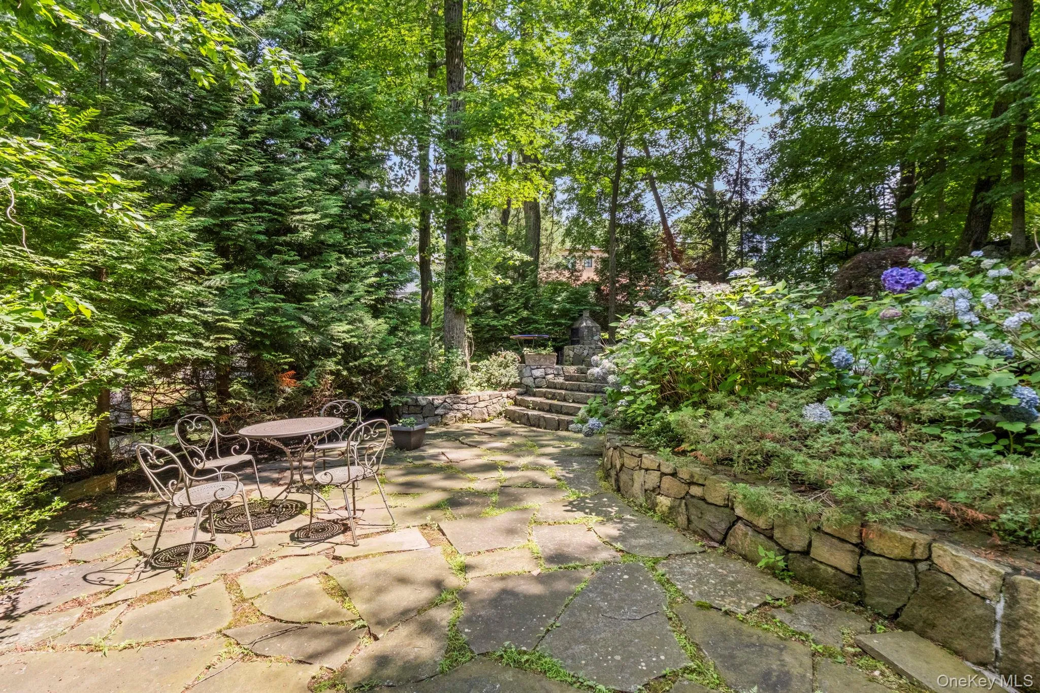 Expansive outdoor patio featuring irregular flagstone paving, a tiered stone retaining wall, and mature landscaping with steps leading upwards Expansive outdoor patio featuring irregular flagstone paving, a tiered stone retaining wall, and mature landscaping with steps leading upwards