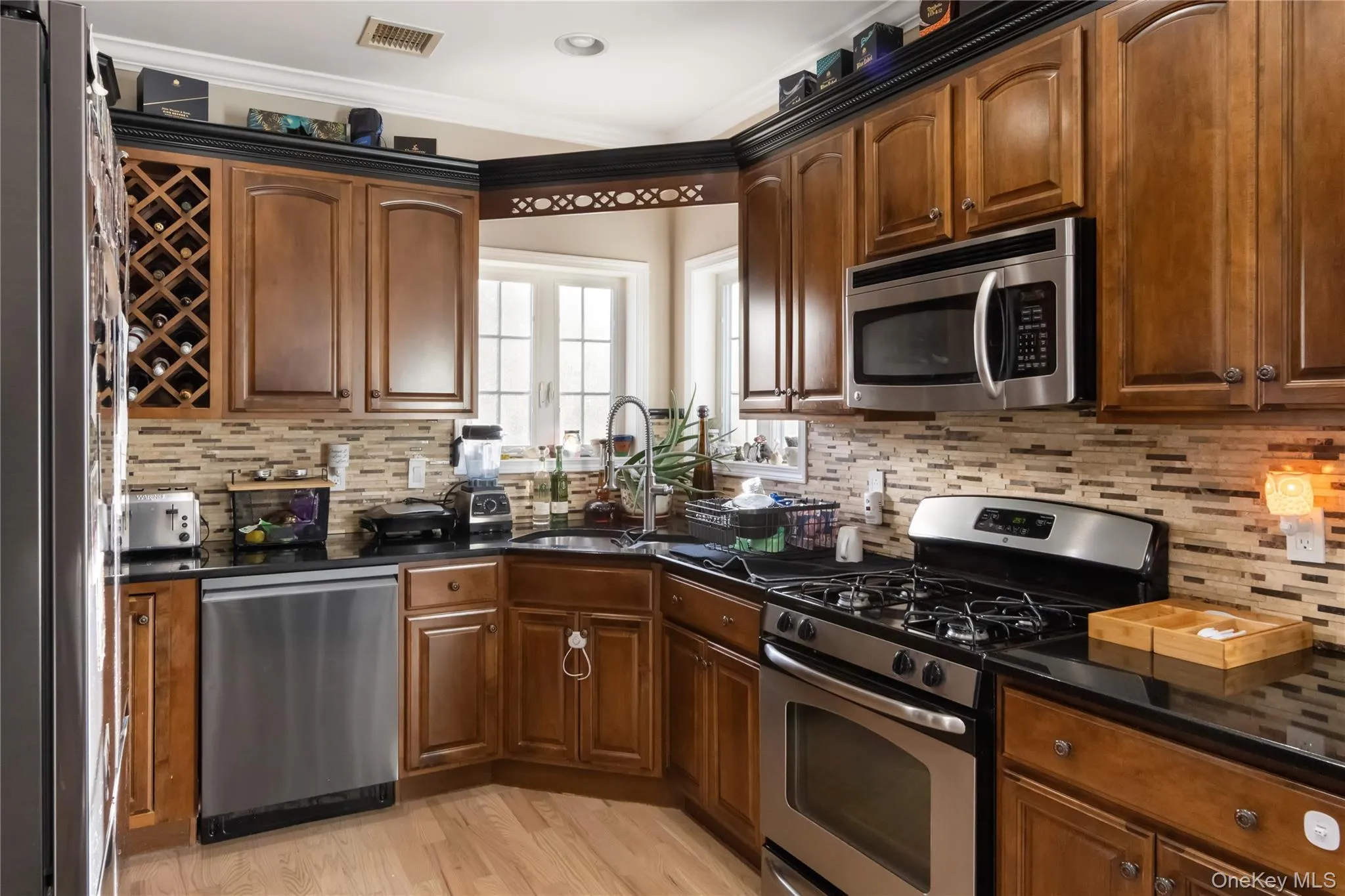 Kitchen featuring stainless steel appliances, light wood-style floors, dark stone counters, ornamental molding, and backsplash Kitchen featuring stainless steel appliances, light wood-style floors, dark stone counters, ornamental molding, and backsplash