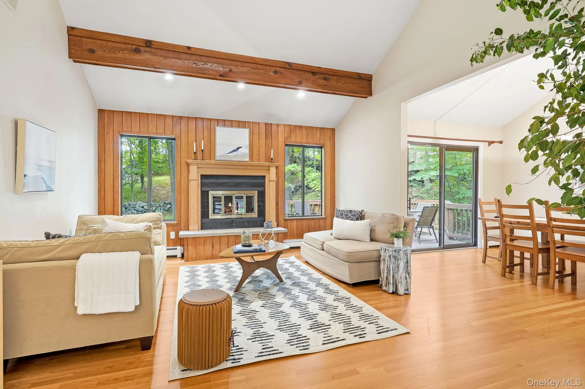 The living room features a wood-burning fireplace with a stone hearth, oversized windows, and a sliding glass door to the exterior deck The living room features a wood-burning fireplace with a stone hearth, oversized windows, and a sliding glass door to the exterior deck