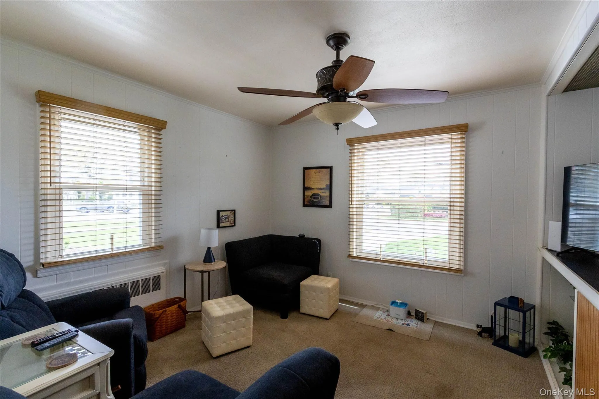 Living area featuring two windows with wood-finish blinds, a ceiling fan with integrated lighting, white vertical paneling on the walls, and carpeted flooring Living area featuring two windows with wood-finish blinds, a ceiling fan with integrated lighting, white vertical paneling on the walls, and carpeted flooring