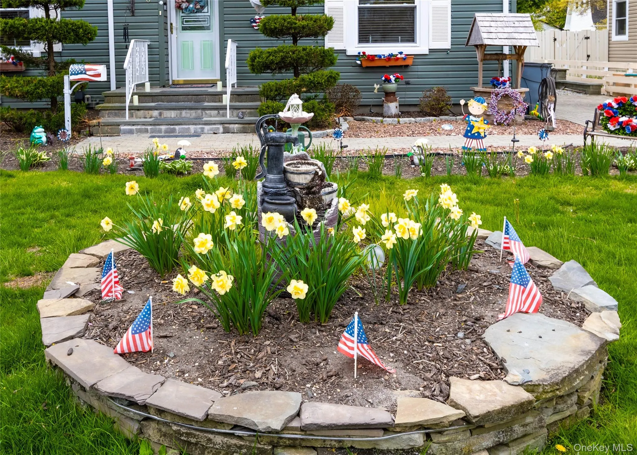 Front yard featuring a stone-lined flower bed with yellow blooms, a manicured lawn, and a paved walkway leading to the entrance Front yard featuring a stone-lined flower bed with yellow blooms, a manicured lawn, and a paved walkway leading to the entrance