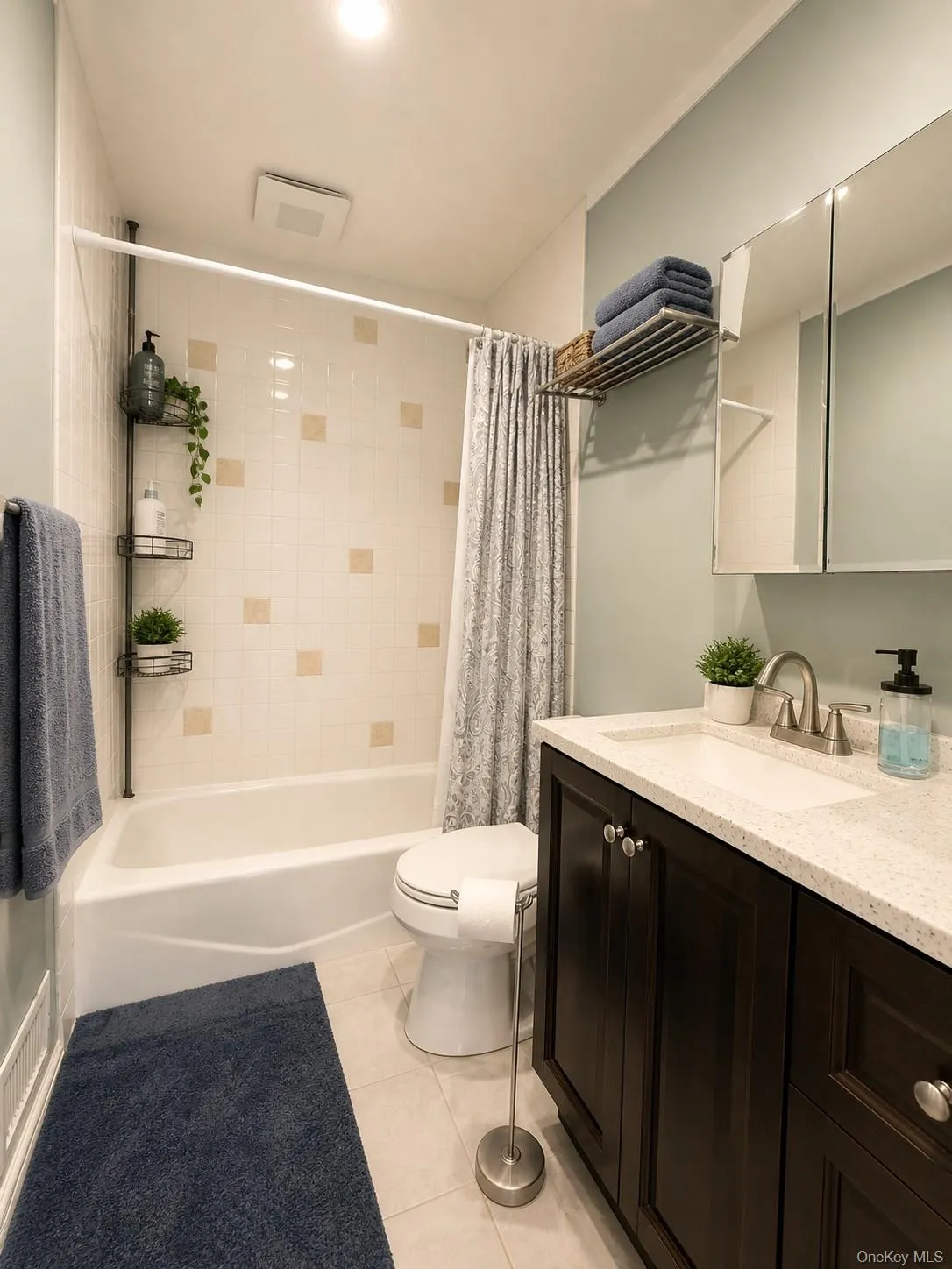Full bathroom featuring a white bathtub with tiled surround, a dark wood-finish vanity with an integrated sink, a speckled light-toned countertop, and tile flooring Full bathroom featuring a white bathtub with tiled surround, a dark wood-finish vanity with an integrated sink, a speckled light-toned countertop, and tile flooring
