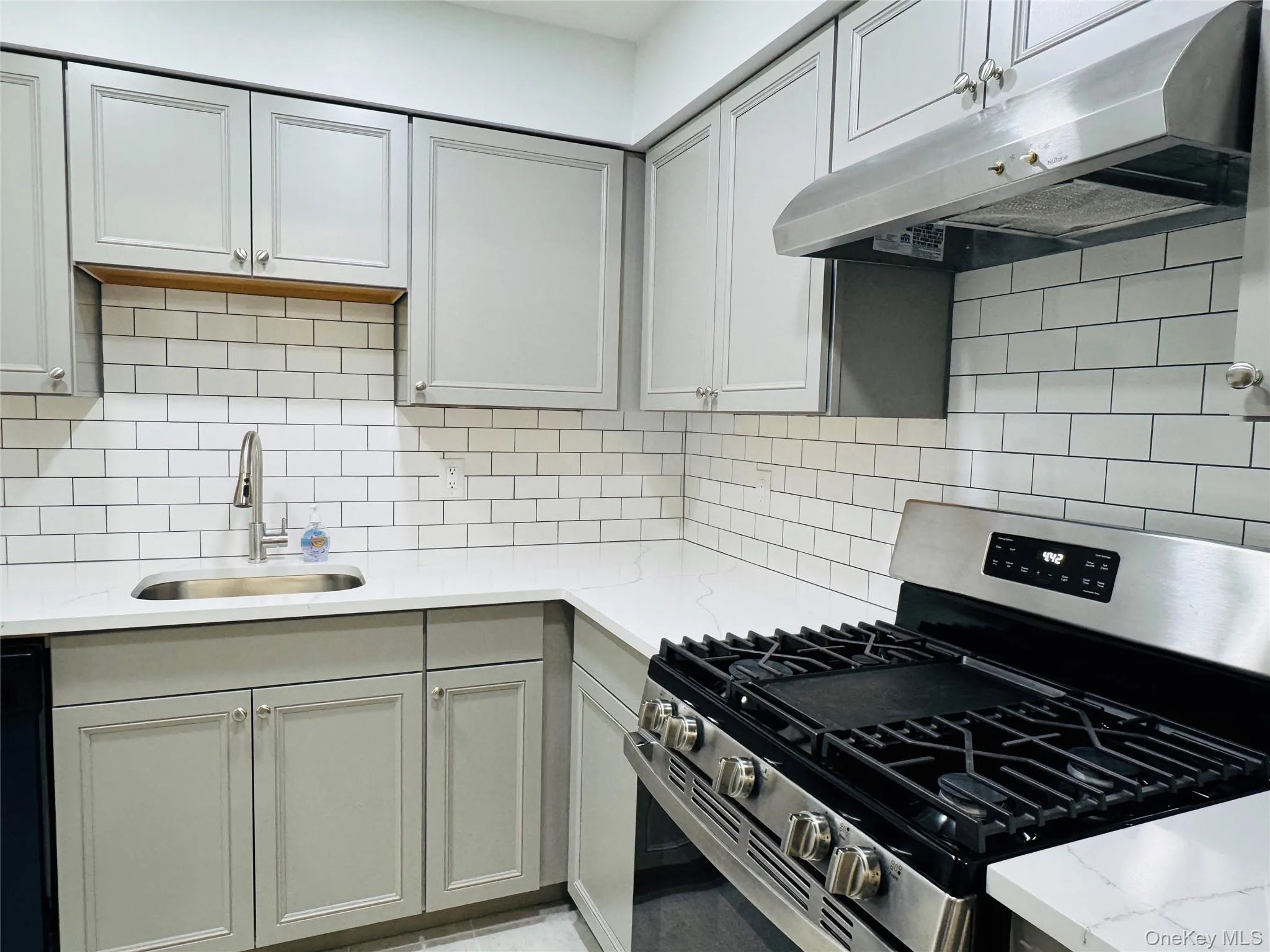 Modern kitchen featuring light gray cabinetry, white subway tile backsplash, white countertops, a stainless steel gas range, and a stainless steel sink with a gooseneck faucet Modern kitchen featuring light gray cabinetry, white subway tile backsplash, white countertops, a stainless steel gas range, and a stainless steel sink with a gooseneck faucet