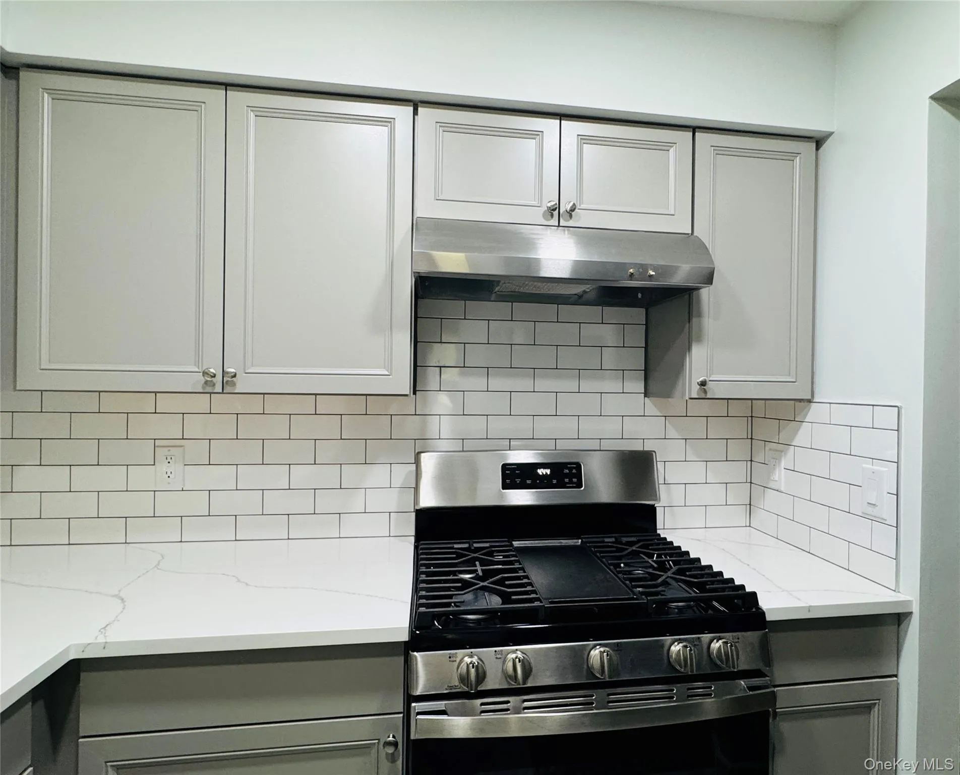 Kitchen featuring light gray cabinetry, a stainless steel range, a coordinating stainless steel range hood, white subway tile backsplash, and light-colored quartz countertops Kitchen featuring light gray cabinetry, a stainless steel range, a coordinating stainless steel range hood, white subway tile backsplash, and light-colored quartz countertops
