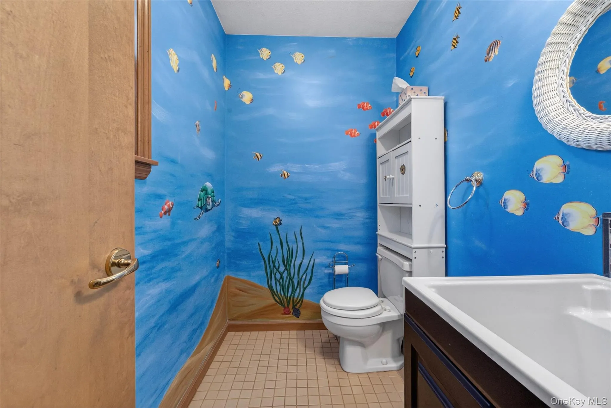 half bath featuring a blue painted wall with aquatic-themed decor, white toilet, white cabinet storage, dark wood-finish vanity with an integrated deep sip sink, and light-colored tile flooring half bath featuring a blue painted wall with aquatic-themed decor, white toilet, white cabinet storage, dark wood-finish vanity with an integrated deep sip sink, and light-colored tile flooring