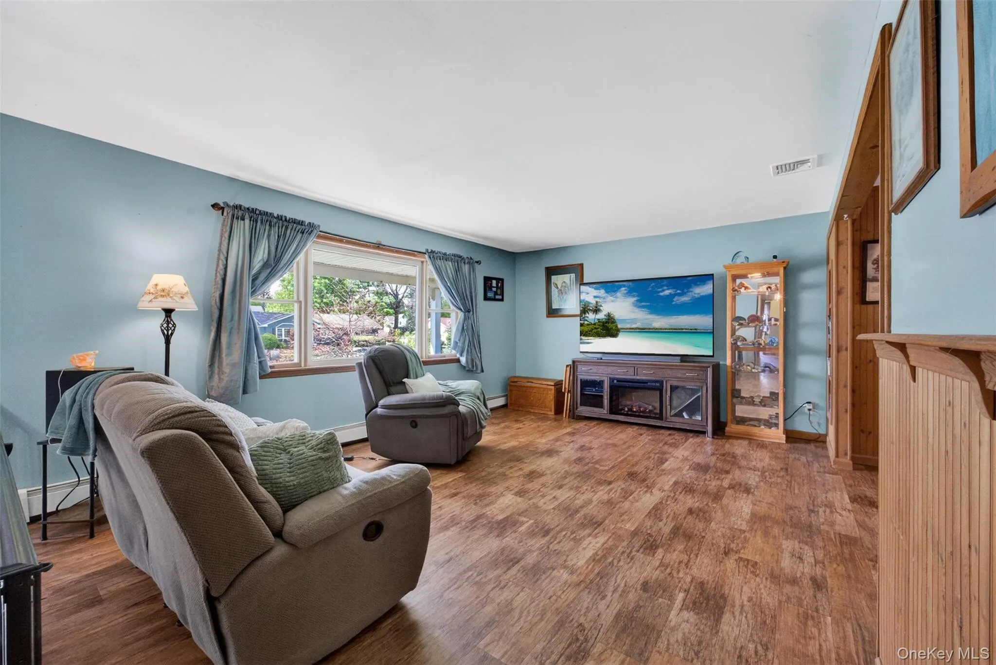Spacious living area featuring wood-finish flooring, light blue walls, and a large window with natural light Spacious living area featuring wood-finish flooring, light blue walls, and a large window with natural light
