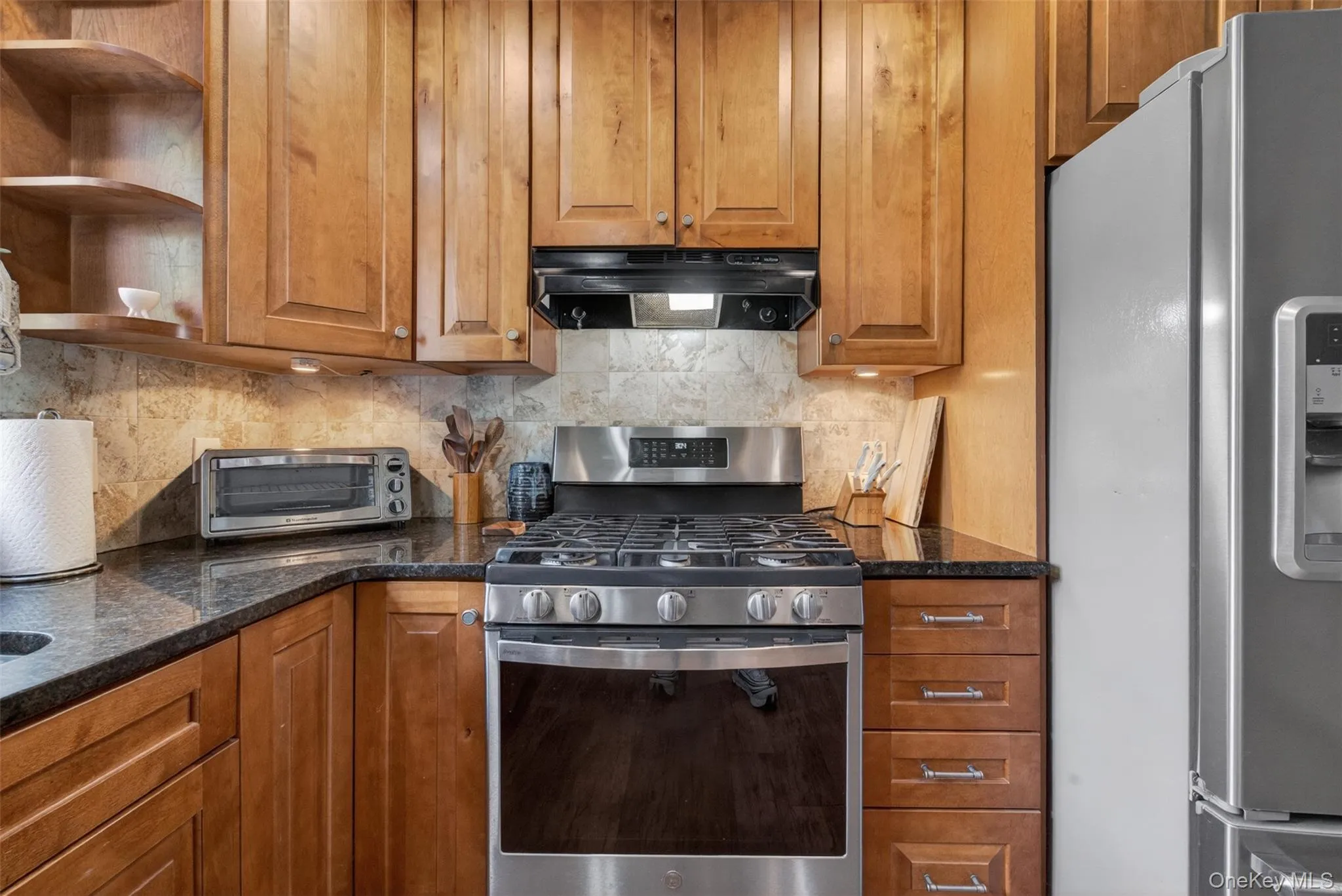 Kitchen featuring a gas range, stainless steel appliances, dark granite countertops, wood cabinetry, and a tiled backsplash Kitchen featuring a gas range, stainless steel appliances, dark granite countertops, wood cabinetry, and a tiled backsplash