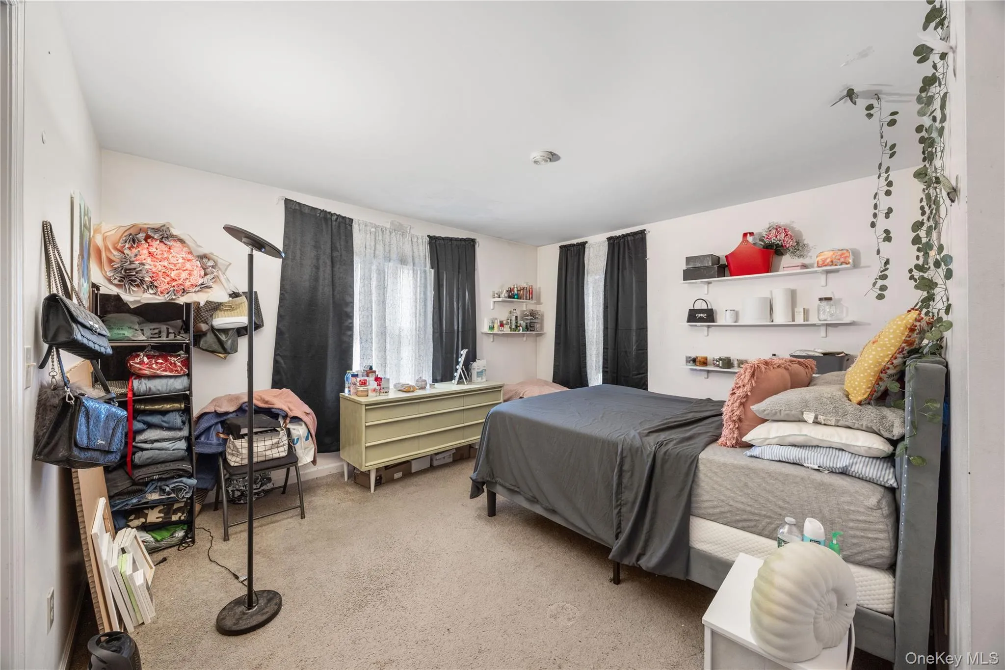 Carpeted room with white walls, two windows featuring dark and sheer curtains, a light green dresser, and built-in wall shelving Carpeted room with white walls, two windows featuring dark and sheer curtains, a light green dresser, and built-in wall shelving