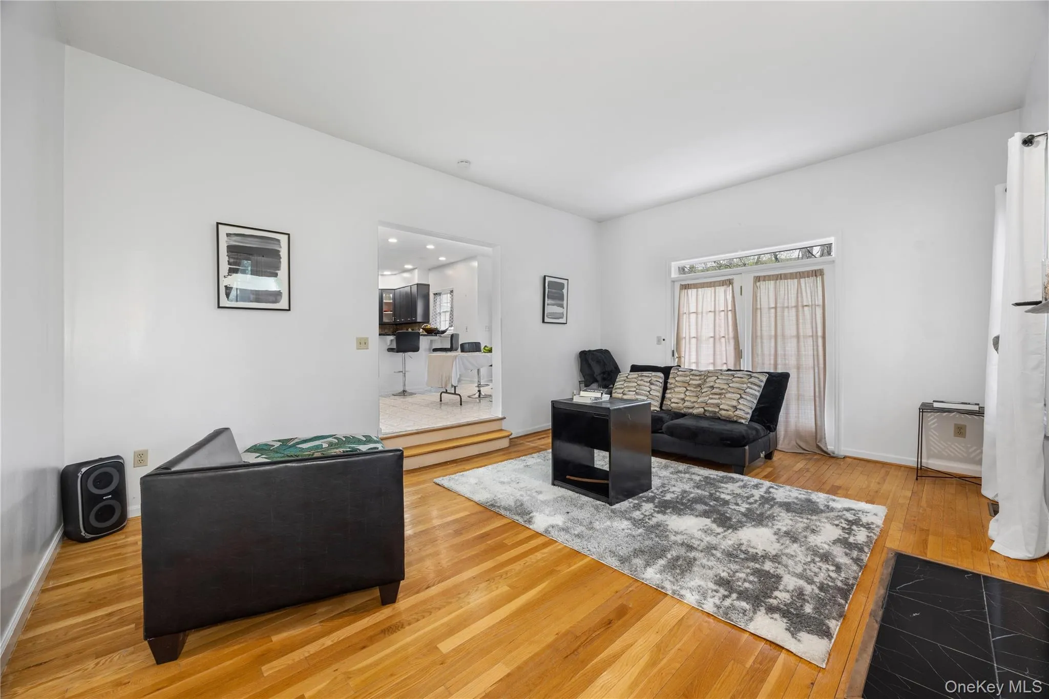 Spacious living area featuring polished wood-finish flooring, white walls, and a black tile hearth Spacious living area featuring polished wood-finish flooring, white walls, and a black tile hearth