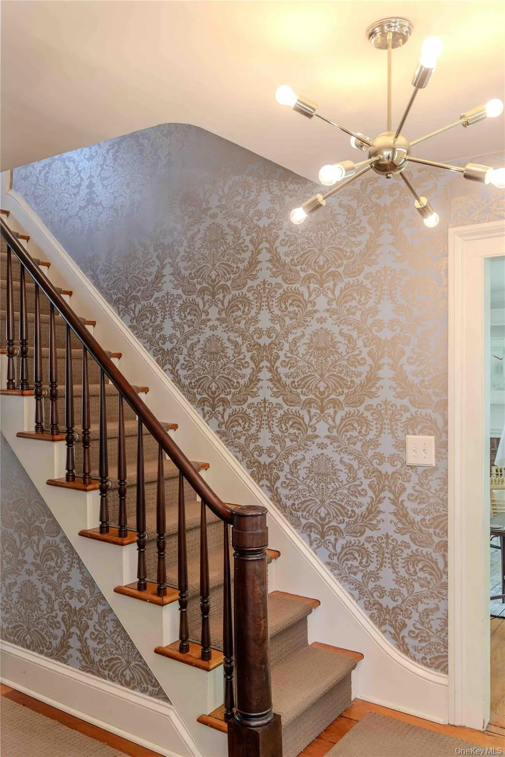 Staircase featuring dark wood newel post and handrail, carpeted treads, and ornate patterned wallpaper Staircase featuring dark wood newel post and handrail, carpeted treads, and ornate patterned wallpaper