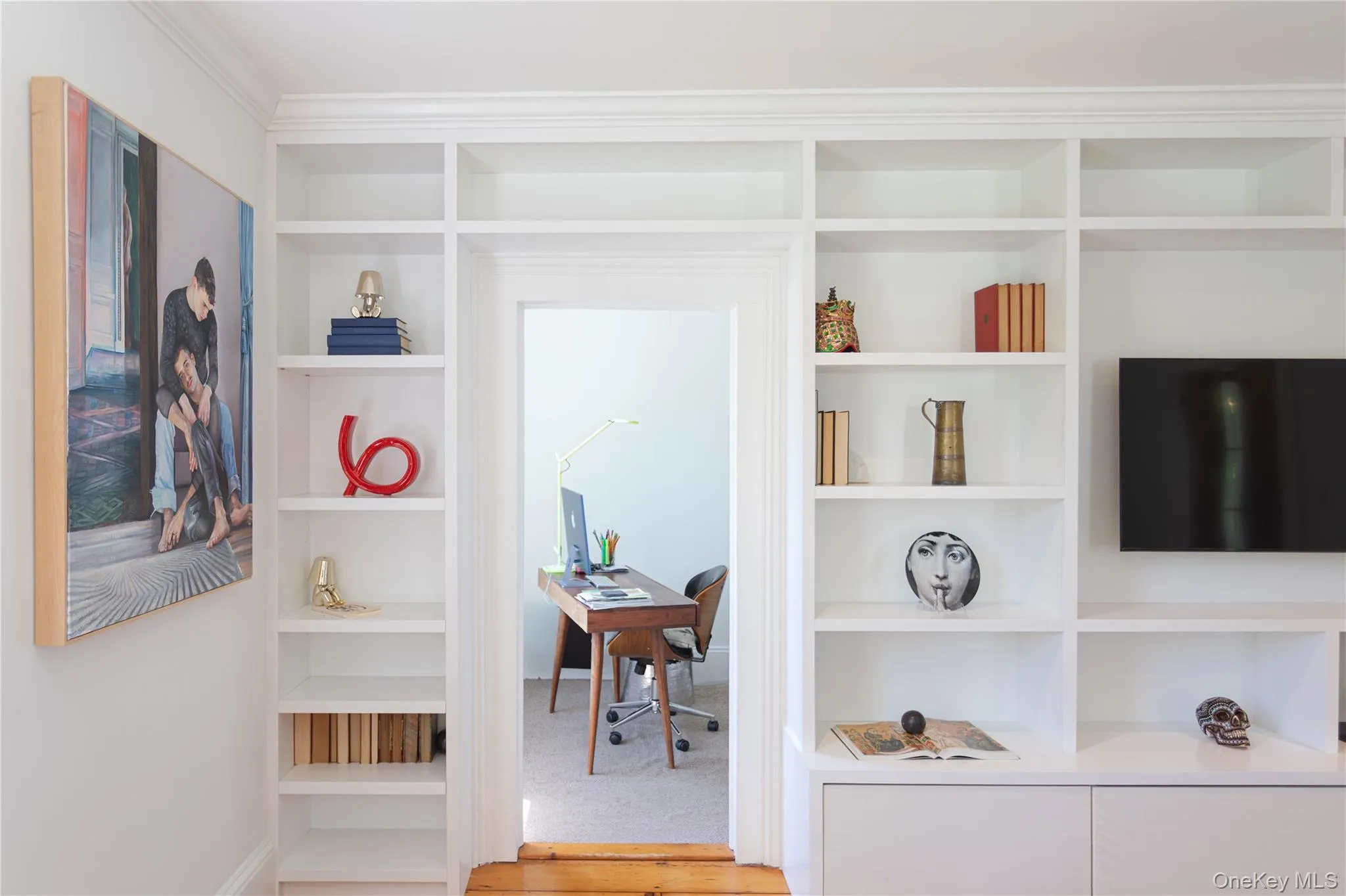 Built-in custom shelving and cabinetry featuring crown molding, an integrated television mount, and a doorway opening to an additional room/studio Built-in custom shelving and cabinetry featuring crown molding, an integrated television mount, and a doorway opening to an additional room/studio