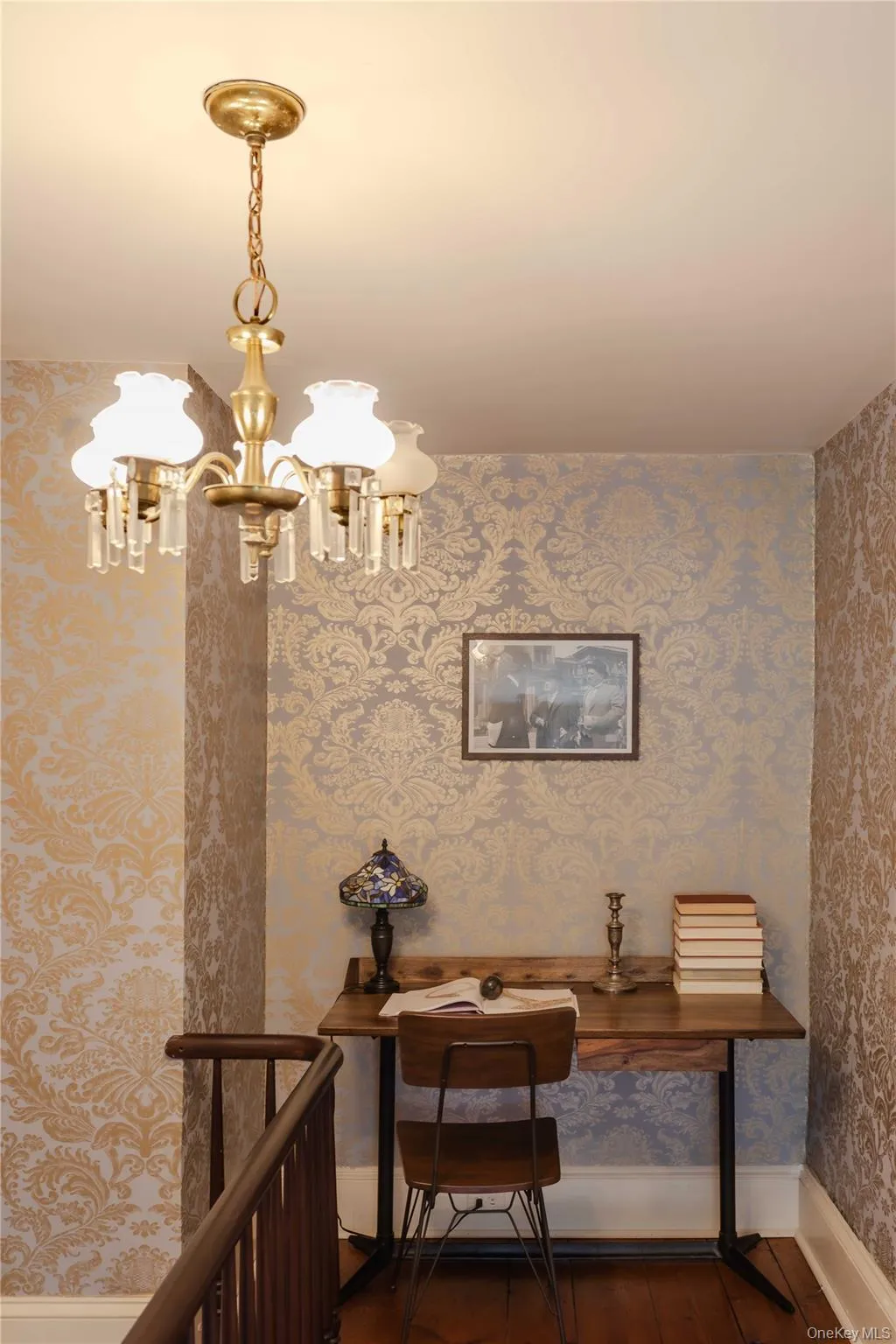 Upper landing featuring patterned wallpaper, wood-finish flooring, and a brass-tone chandelier with crystal accents Upper landing featuring patterned wallpaper, wood-finish flooring, and a brass-tone chandelier with crystal accents