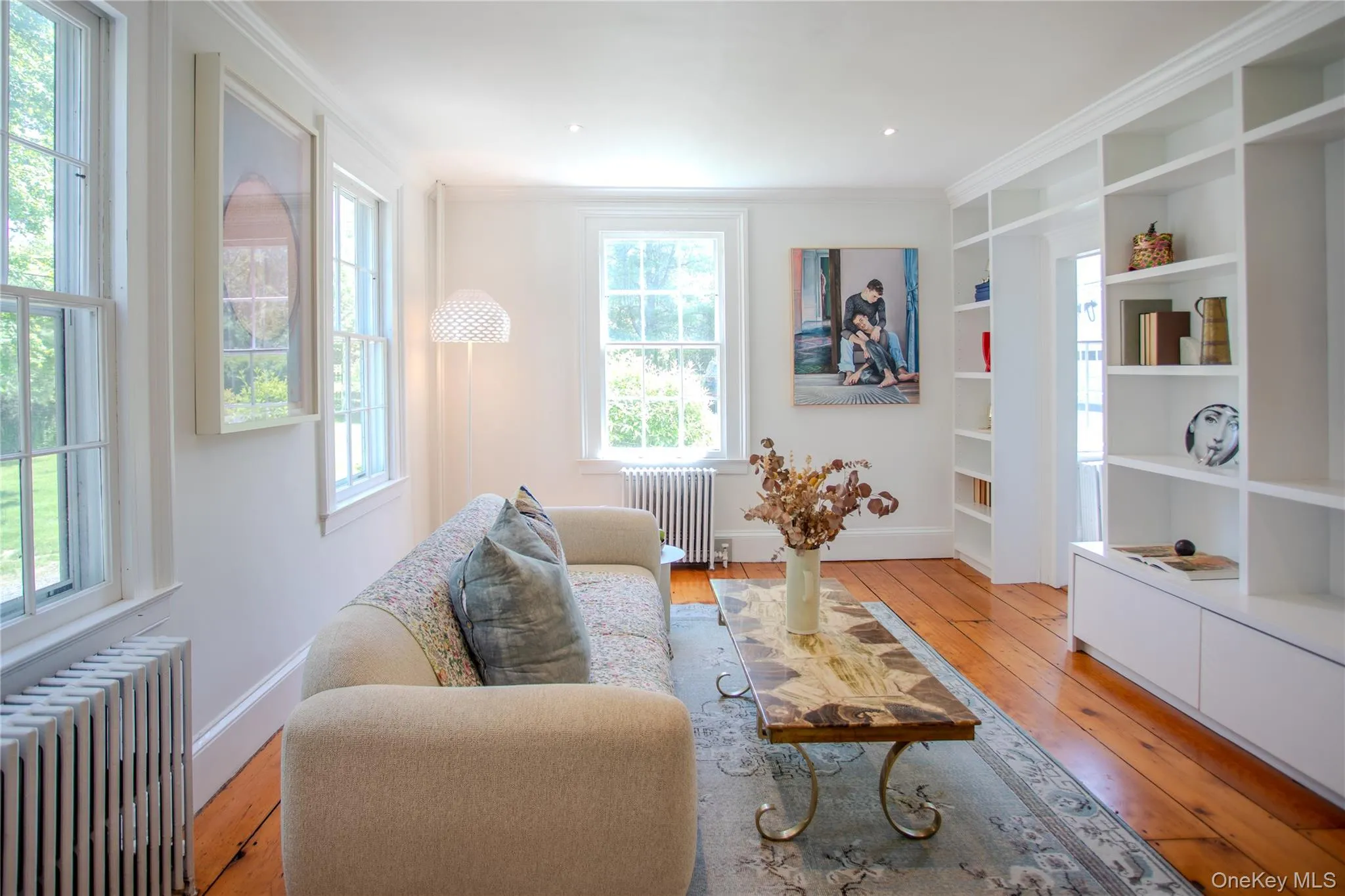Light-filled living space featuring multiple large windows, wide plank wood flooring, extensive built-in shelving Light-filled living space featuring multiple large windows, wide plank wood flooring, extensive built-in shelving