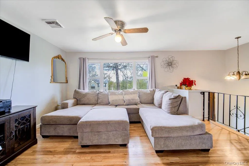 Spacious living area featuring wood-finish flooring, a large window, a ceiling fan with integrated lighting, and a decorative wrought iron railing Spacious living area featuring wood-finish flooring, a large window, a ceiling fan with integrated lighting, and a decorative wrought iron railing