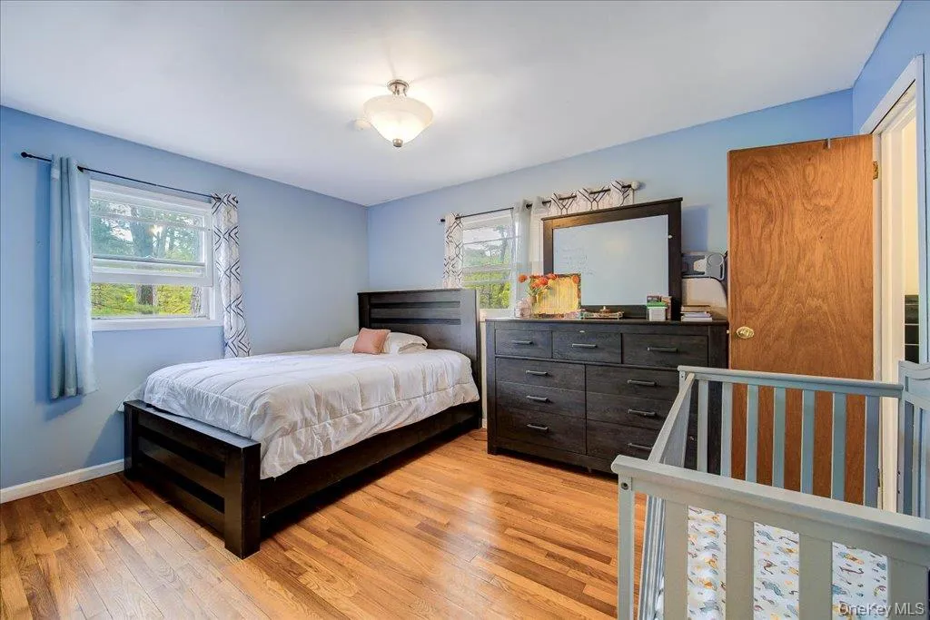 Primary bedroom with Hardwood-finish flooring featuring a flush-mount ceiling light fixture, two windows with white trim, and a solid wood door Primary bedroom with Hardwood-finish flooring featuring a flush-mount ceiling light fixture, two windows with white trim, and a solid wood door