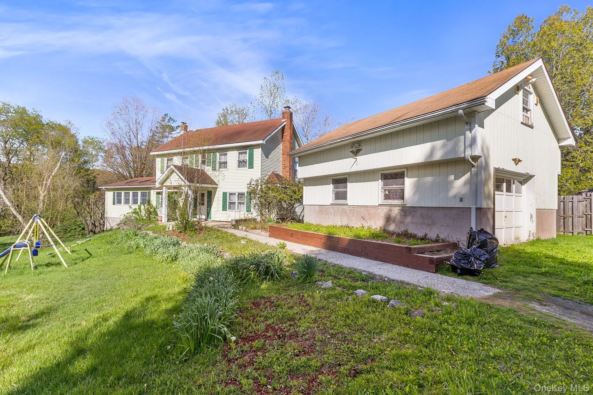 Light-colored siding home with green accent shutters, a front porch, and an attached two-story structure featuring a garage door Light-colored siding home with green accent shutters, a front porch, and an attached two-story structure featuring a garage door