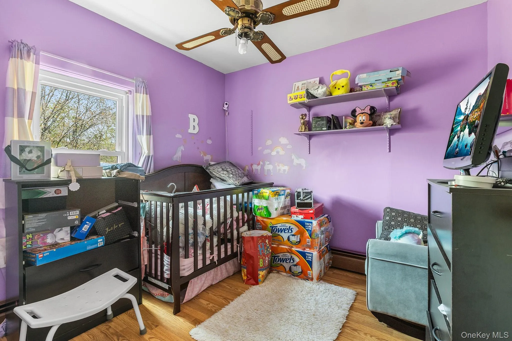 Hardwood-finish flooring complements the purple wall finish in this room featuring a window with a white frame, a ceiling fan with wood-finish blades, and wall-mounted shelving Hardwood-finish flooring complements the purple wall finish in this room featuring a window with a white frame, a ceiling fan with wood-finish blades, and wall-mounted shelving
