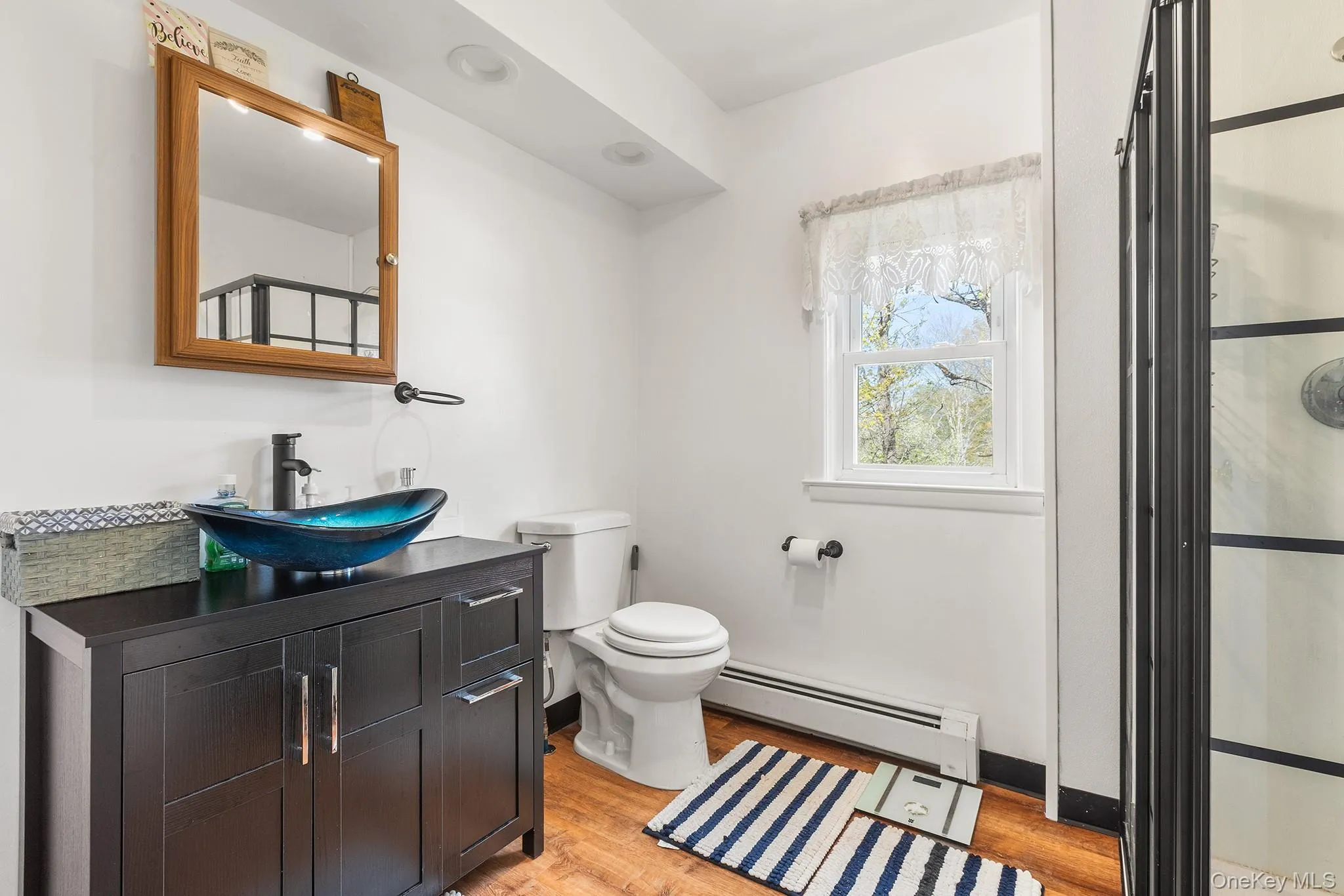 Bathroom featuring a black vanity with a vessel sink, a framed mirror, wood-finish flooring, a window, and a glass-enclosed shower Bathroom featuring a black vanity with a vessel sink, a framed mirror, wood-finish flooring, a window, and a glass-enclosed shower